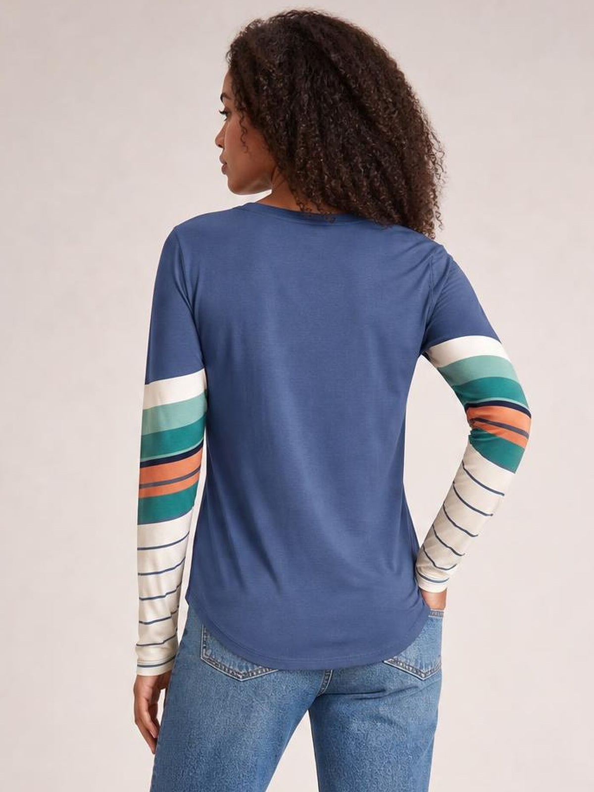 Model viewed from the back wears a blue geometric round-neck long-sleeve T-shirt with multicolor striped sleeves and a relaxed silhouette.