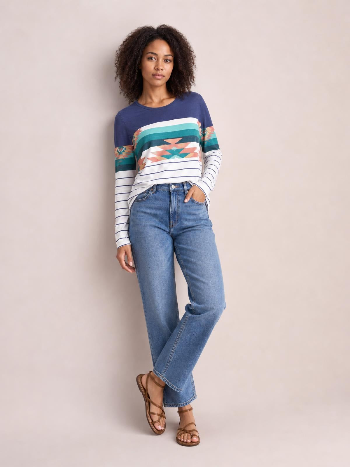 Model in a relaxed crossed-leg pose wears a geometric round neck long sleeve T-shirt with a navy, teal, coral, and white patterned striped design.