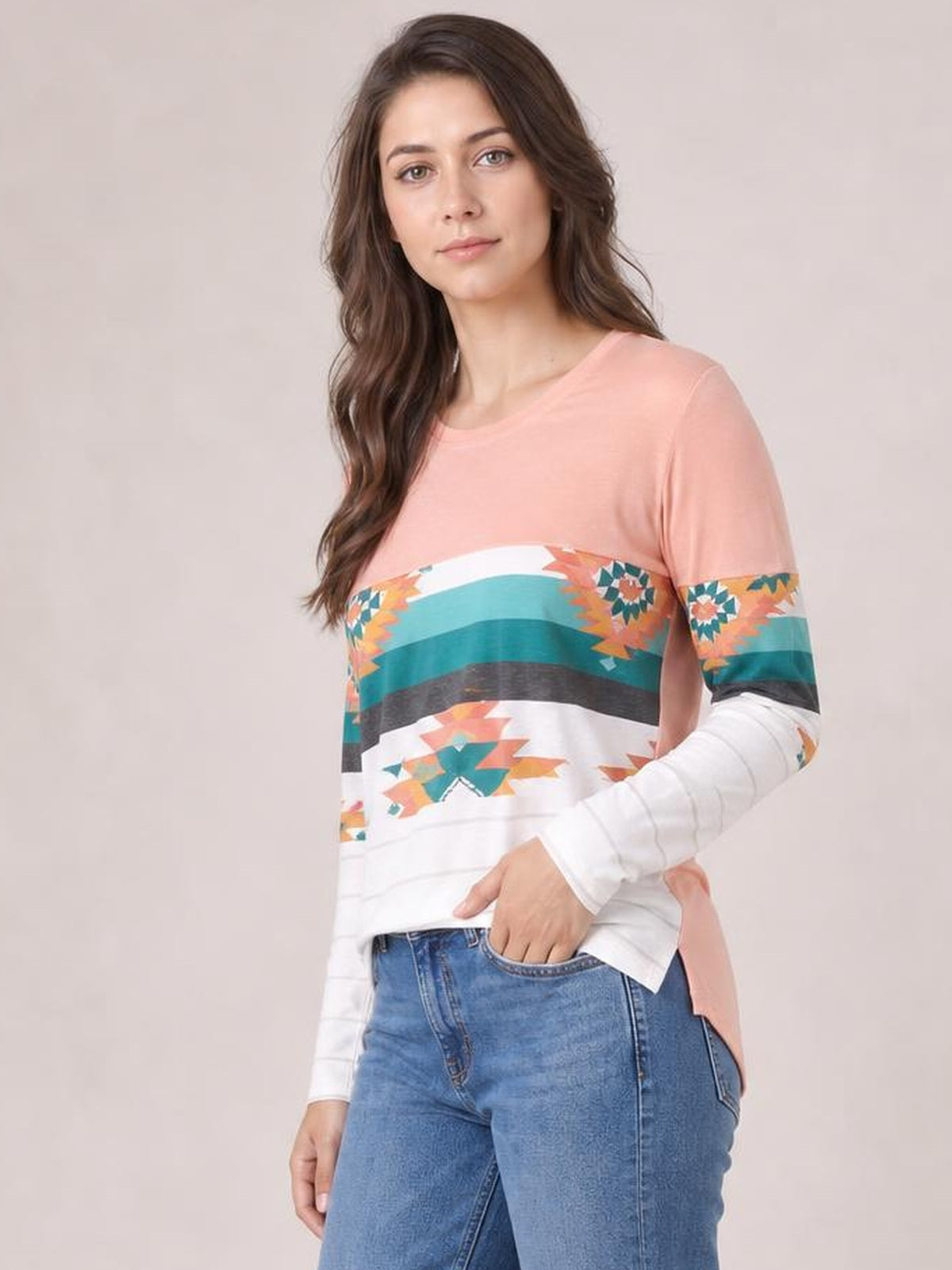 Model wears a pink geometric round neck long sleeve T-shirt with multicolor patterned stripes, an opaque relaxed silhouette, and one hand in her jeans pocket.