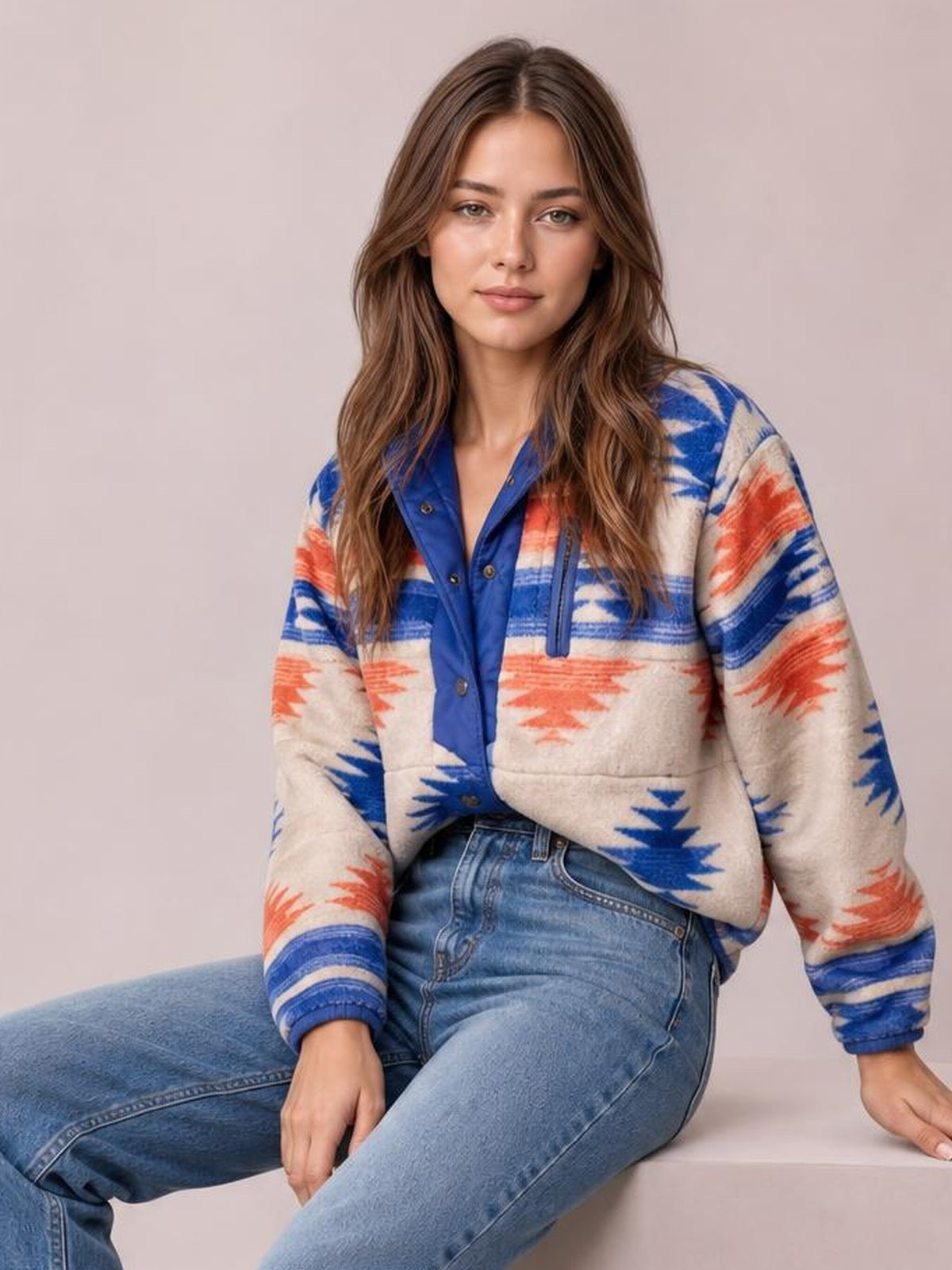 Seated model wears a cream geometric snap-down jacket with blue and orange patterns, button front, long sleeves, ribbed cuffs, and a relaxed silhouette.