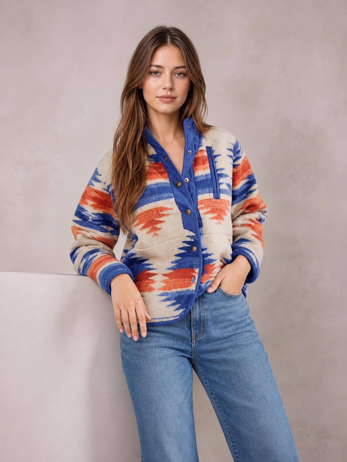 A model stands with one hand in her jeans pocket, wearing a cream geometric snap-down jacket with blue trim, orange and blue patterning, and front pockets.