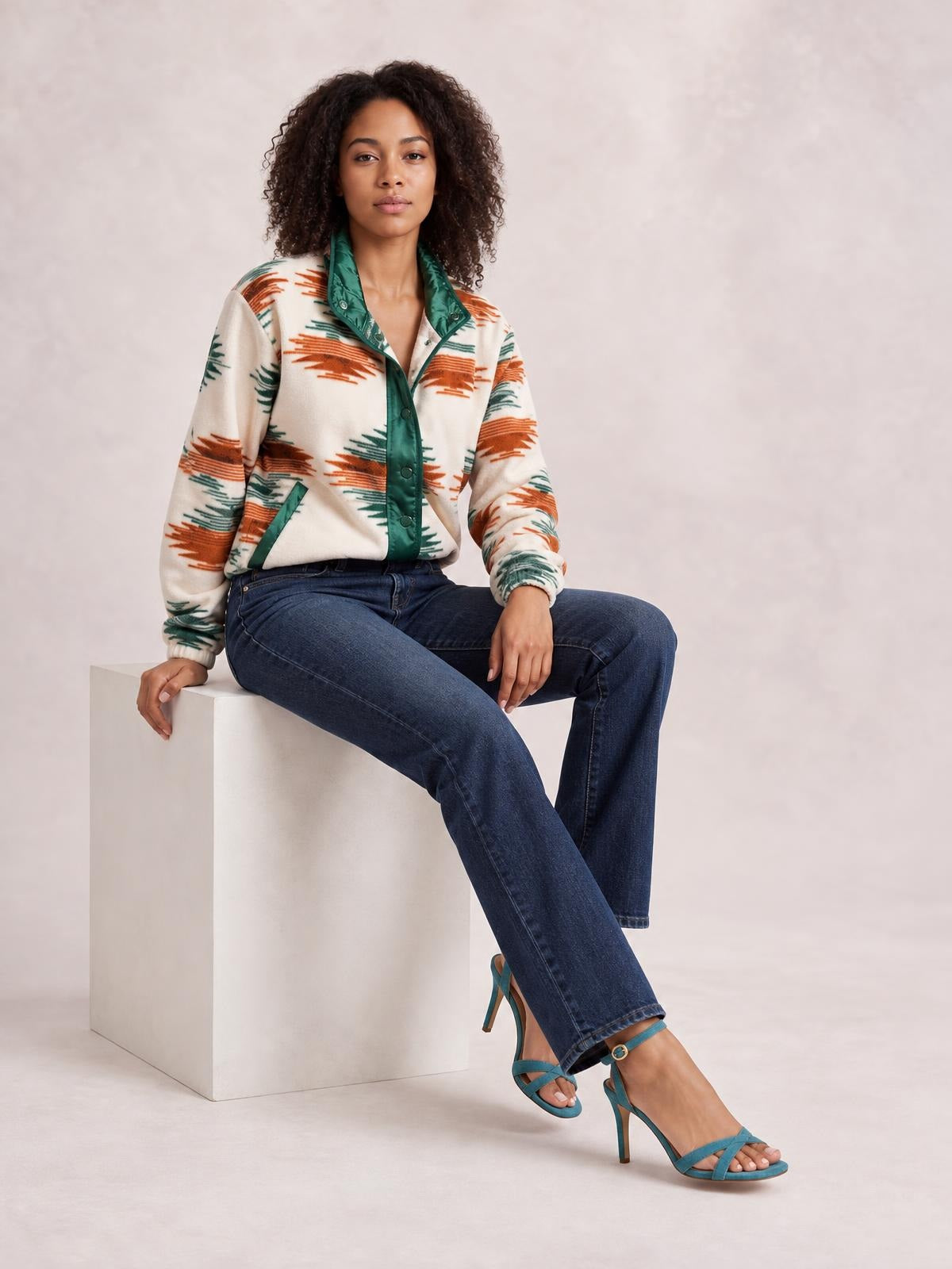 A woman sits wearing a cream Geometric Snap Down Jacket with rust and green geometric print, green snap placket and trim, long sleeves, and a relaxed silhouette.