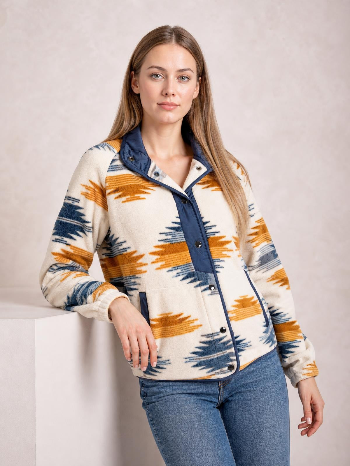 Model leans with one hand on a ledge wearing a cream geometric snap-down jacket with navy trim, long sleeves, snap front, and pockets.