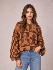 A model wears a brown and tan Gingerbread Man sweater with a round neck, drop shoulders, oversized fit, and voluminous sleeves, posing with one hand in her jeans pocket.