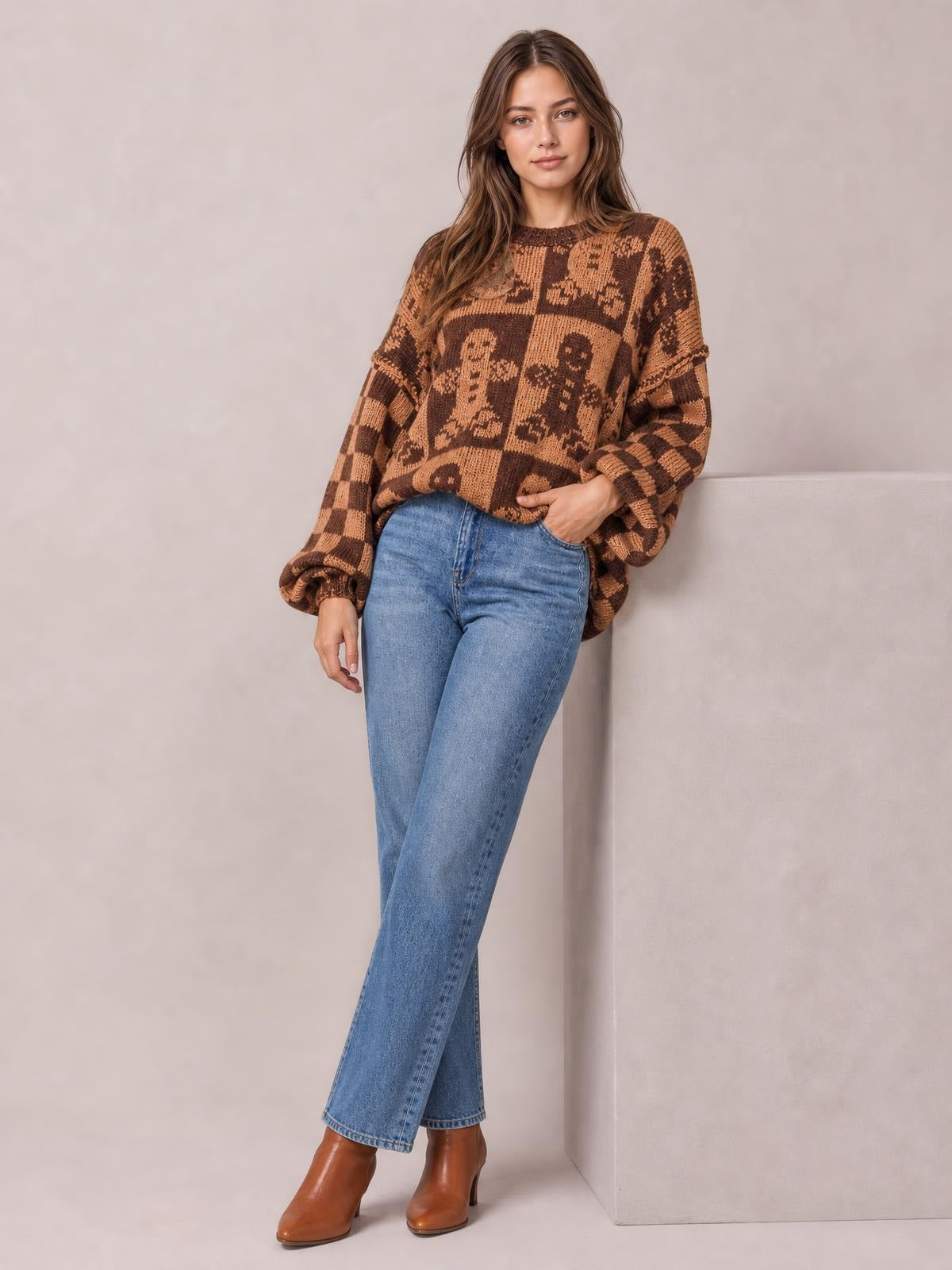 A model poses with one hand in her jeans pocket, wearing a brown-and-tan gingerbread man sweater with a round neck, drop shoulders, relaxed fit, and ribbed cuffs.