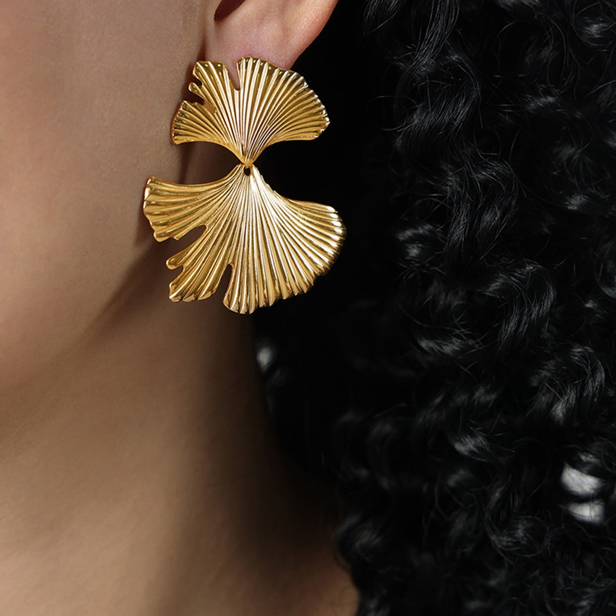 A close-up shows a gold-plated Ginkgo Biloba stud earring with two fan-shaped leaves, fine radial ridges, and a sculptural connected silhouette beside dark hair.