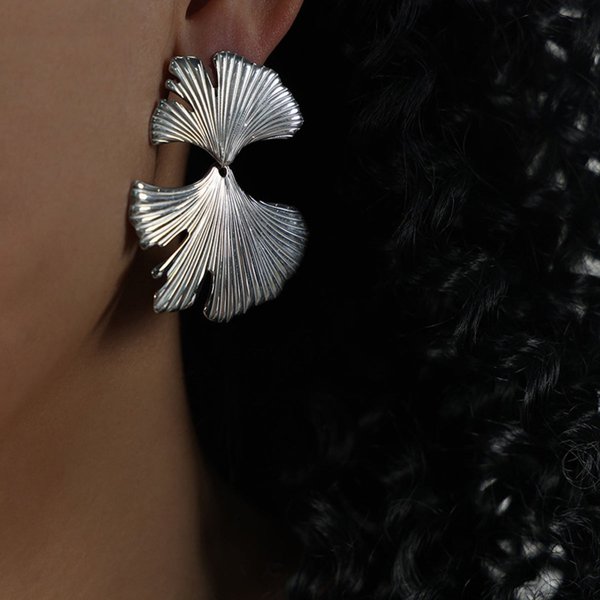 A close-up of a model’s ear shows a metallic Ginkgo Biloba design stud earring with sculpted, ribbed leaf fans and an elongated botanical silhouette.