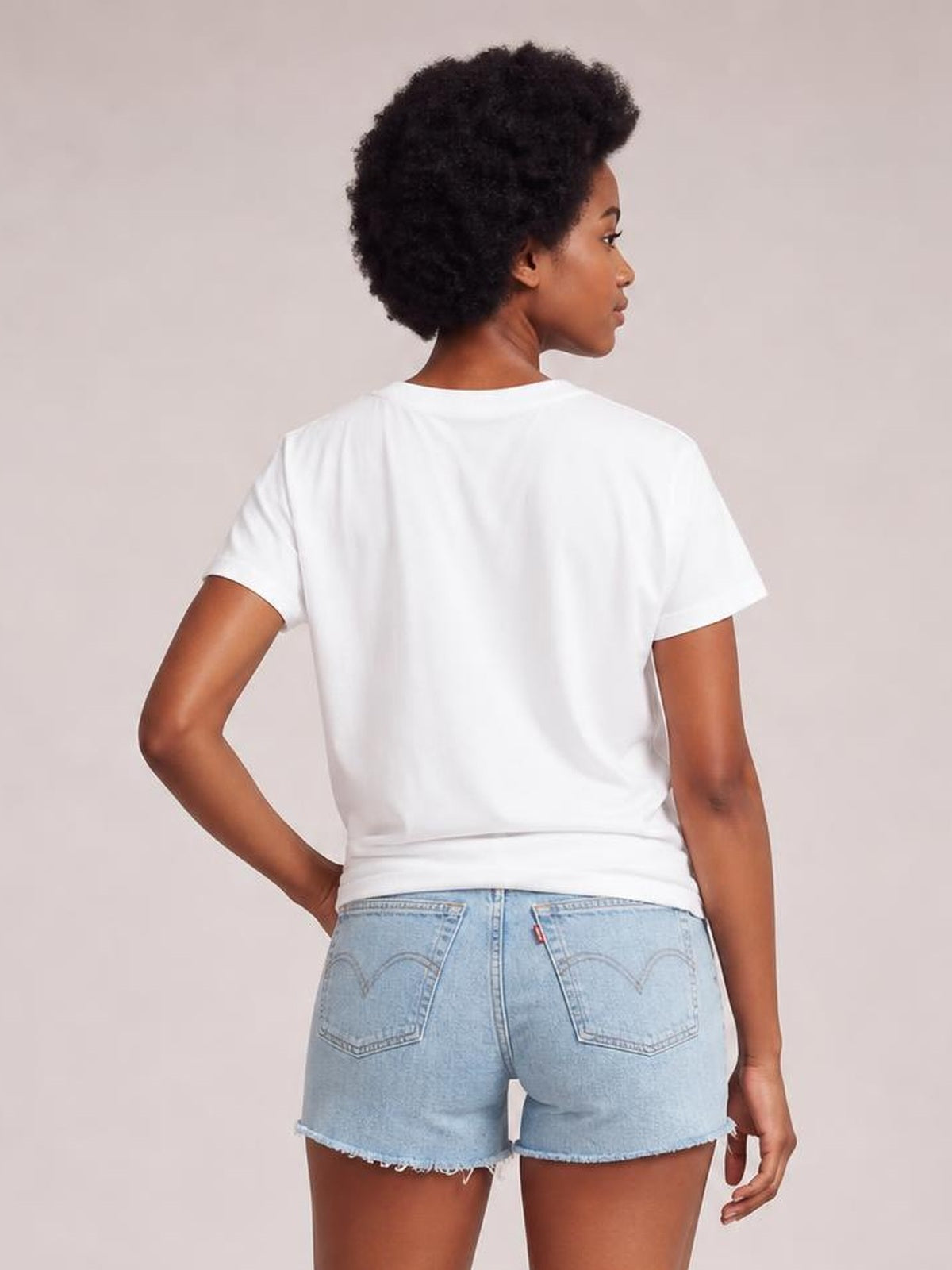 A model poses with one hand on her hip, showing the back of a white graphic round-neck short-sleeve T-shirt with a relaxed silhouette.