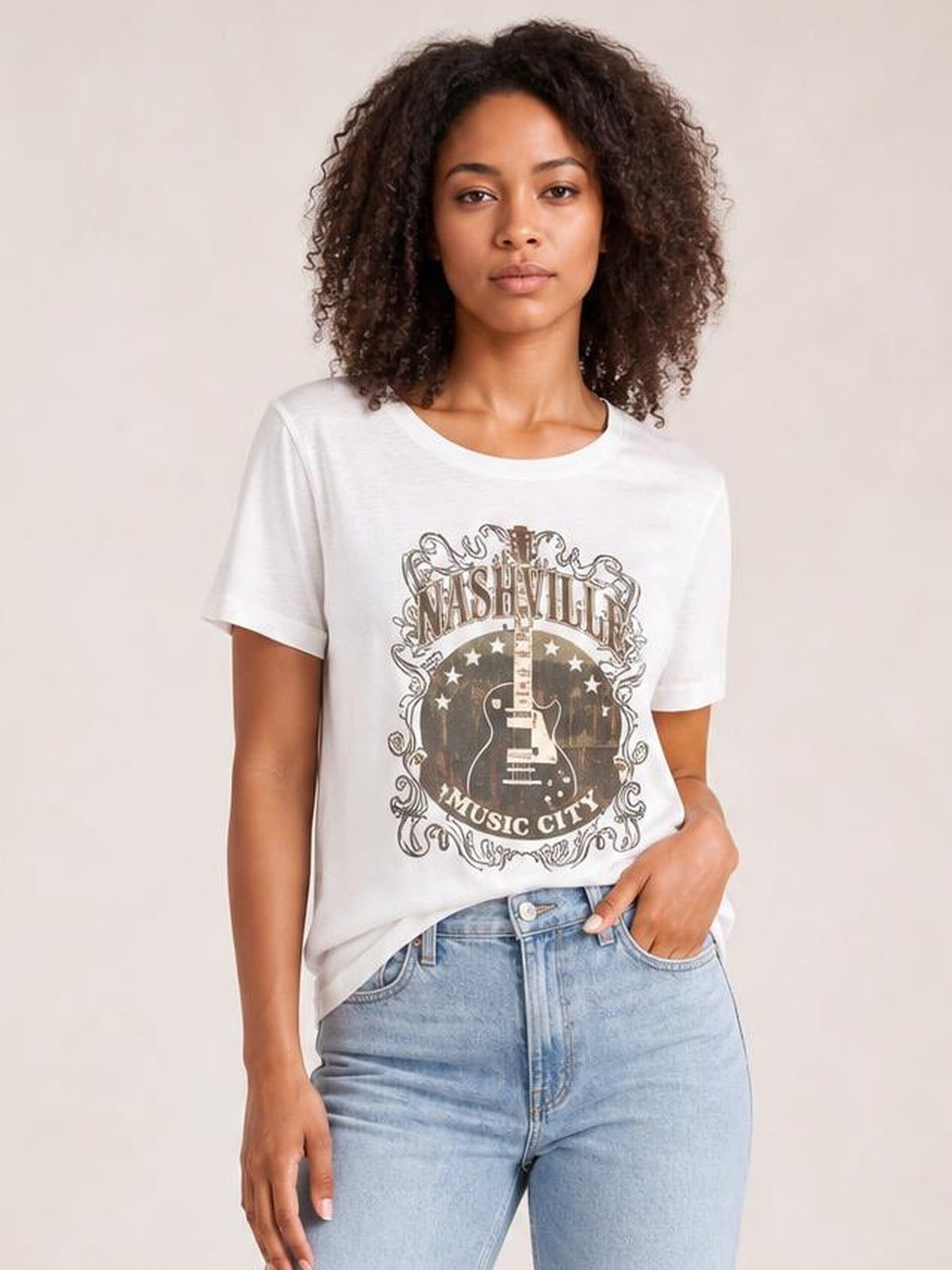 Model wearing an opaque white round neck short sleeve T-shirt with a Nashville guitar graphic, styled in a relaxed fit with one hand in her jeans pocket.