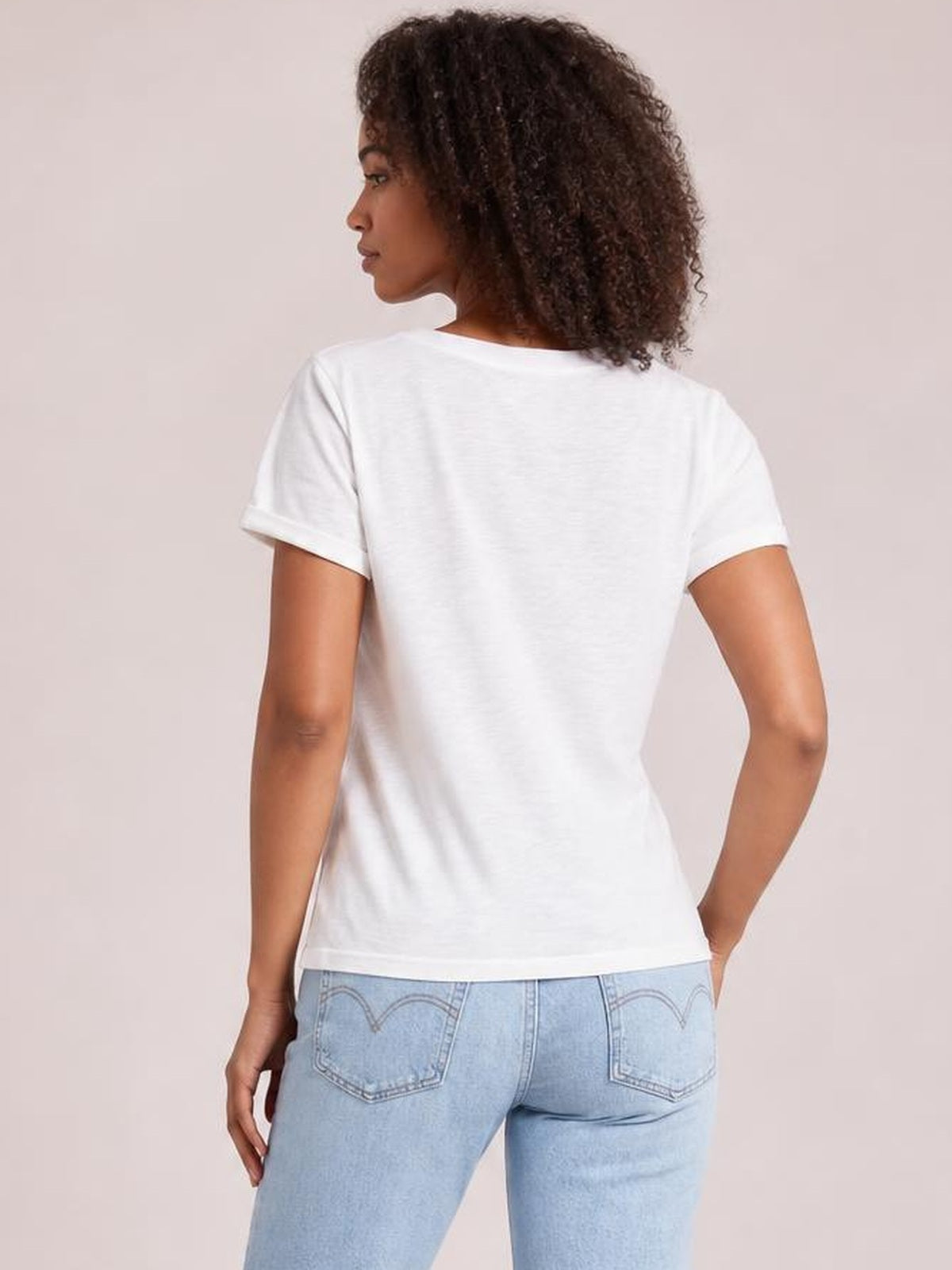 A model looks over her shoulder, showing the back of a white round neck short sleeve T-shirt with a casual, straight silhouette and rolled sleeve hems.