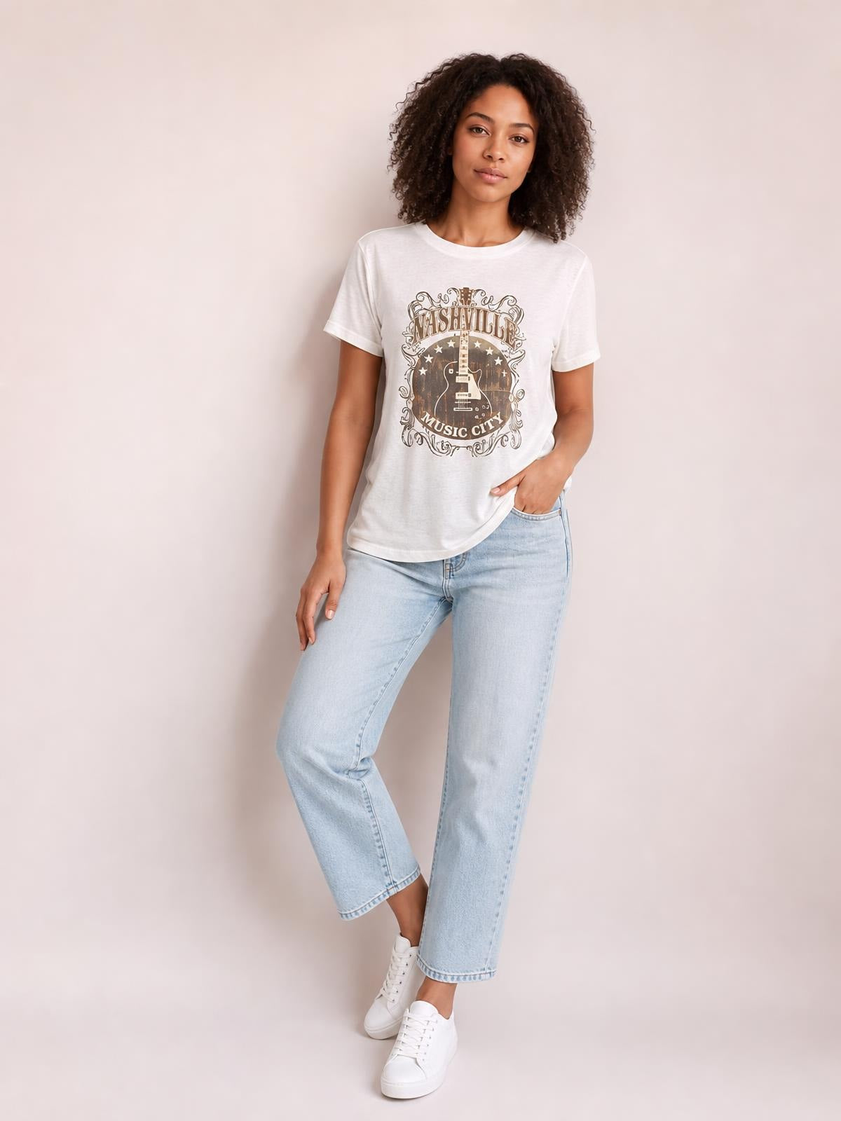 Model wears a white round-neck short-sleeve T-shirt with a vintage guitar graphic, relaxed straight silhouette, and one hand tucked into her jeans pocket.