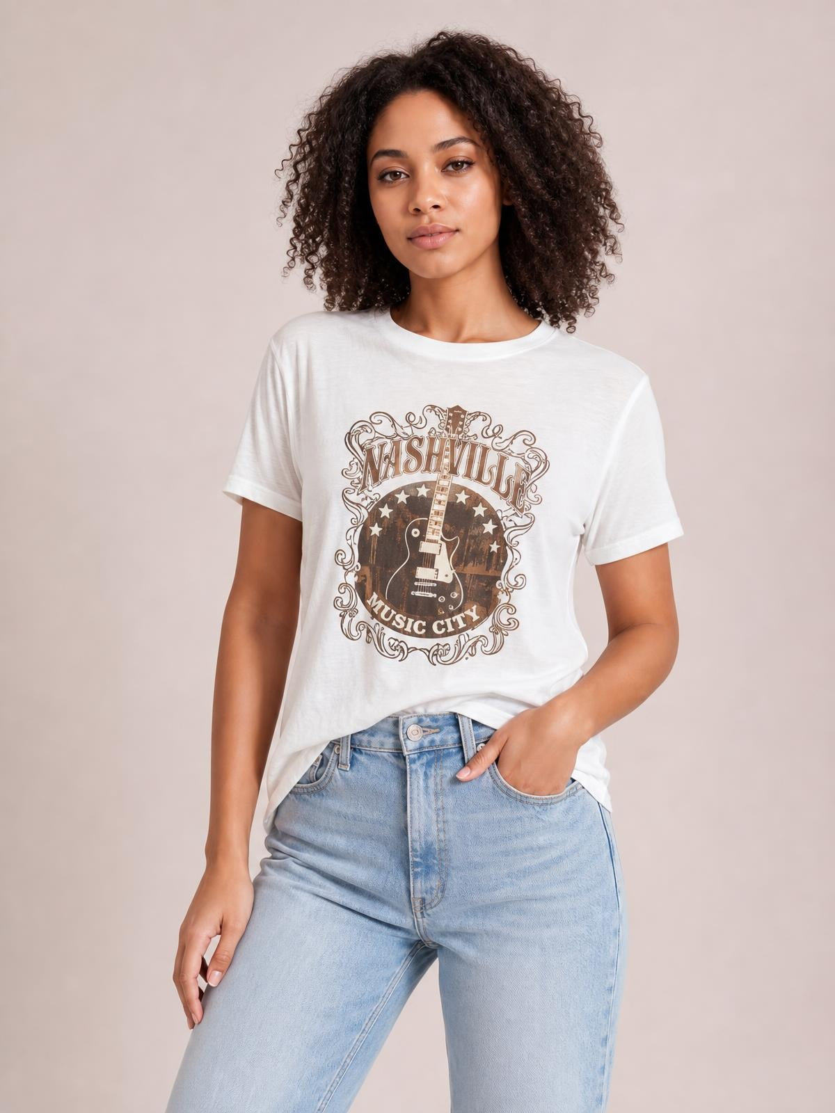 Model poses in a white round-neck short-sleeve T-shirt with a Nashville guitar graphic and relaxed silhouette, one hand in her jeans pocket.