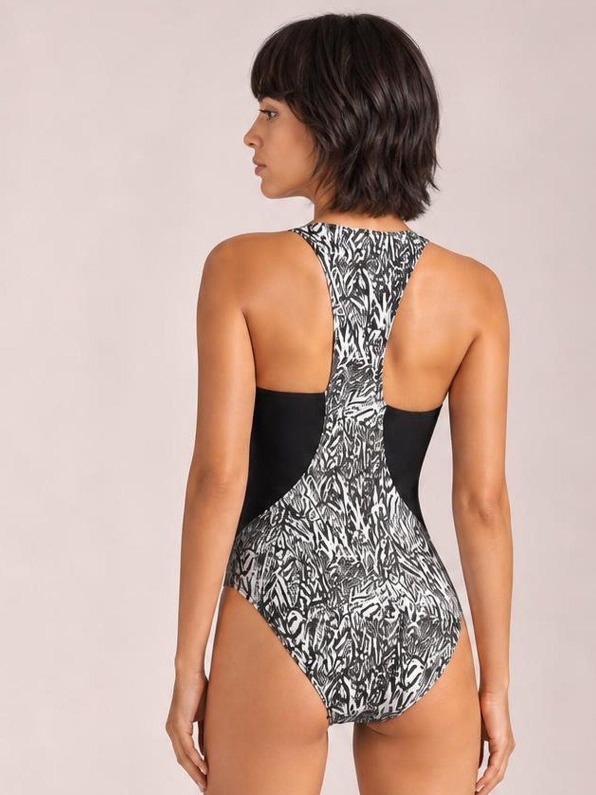 Model shown from behind wearing a black-and-white printed one-piece swimsuit with a high-cut leg, racerback design, and contrasting black side panels.