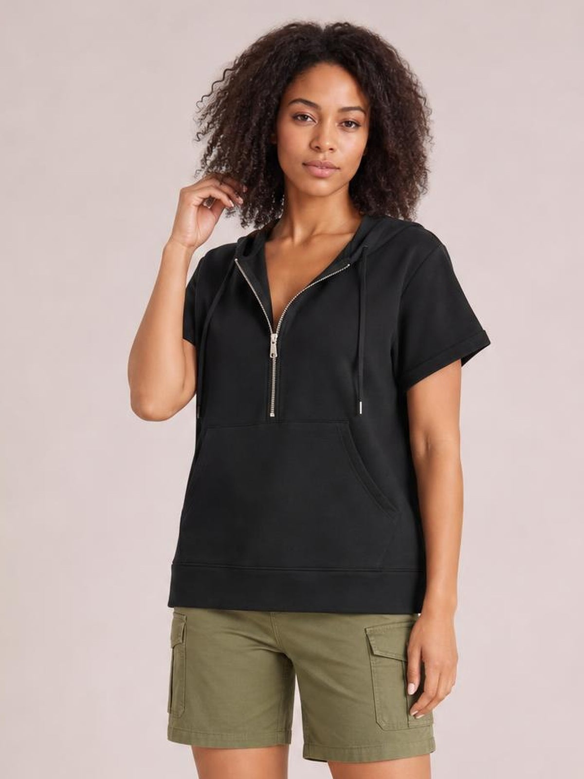 Model poses in a black short-sleeve hooded top with a half zip, relaxed silhouette, drawstring hood, and spacious kangaroo pocket.
