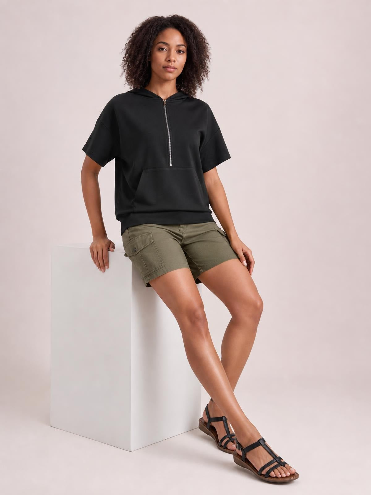 Model seated in a relaxed black short-sleeve hooded top with a half-zip neckline, loose silhouette, and large front kangaroo pocket.