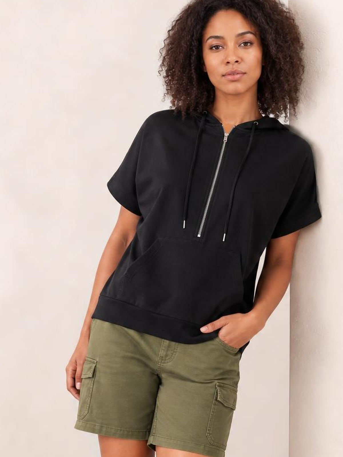 A model leans casually wearing a black short-sleeve hooded top with a half-zip front, drawstrings, roomy pocket, and relaxed silhouette.