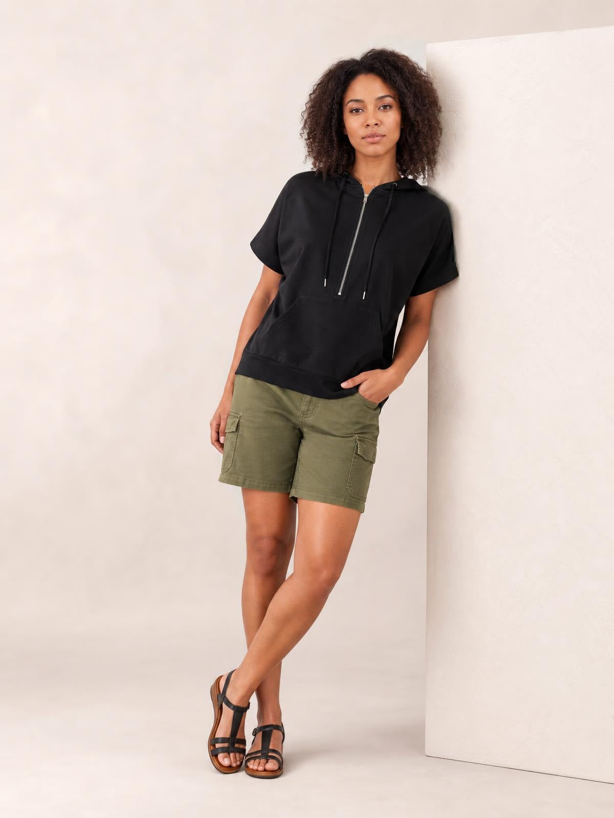 A model leans in a black half-zip short-sleeve hooded top with drawstrings, a relaxed silhouette, and a spacious front kangaroo pocket.