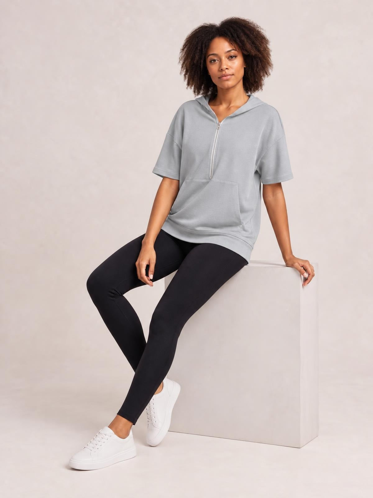 A woman sits in a gray short-sleeve hooded top with a half-zip front, relaxed silhouette, and spacious kangaroo pocket.
