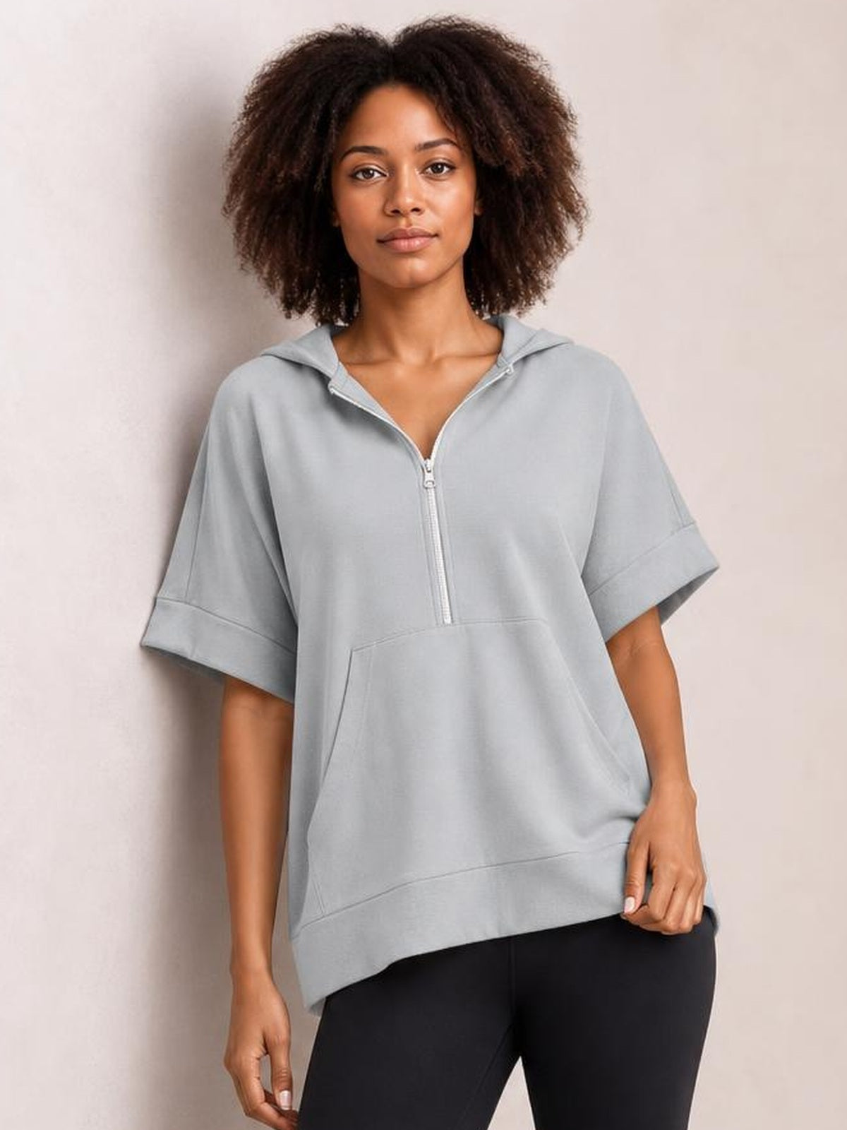 Model wears a light gray half-zip short sleeve hooded top with rolled cuffs, relaxed oversized silhouette, and a large front kangaroo pocket.