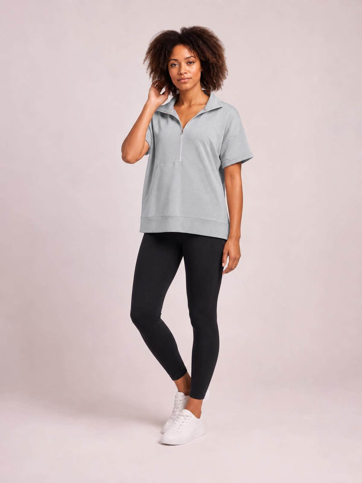 Model wears a light gray short-sleeve half-zip hooded top with a relaxed silhouette, folded cuffs, front pockets, and a straight hem while posing with one leg crossed.