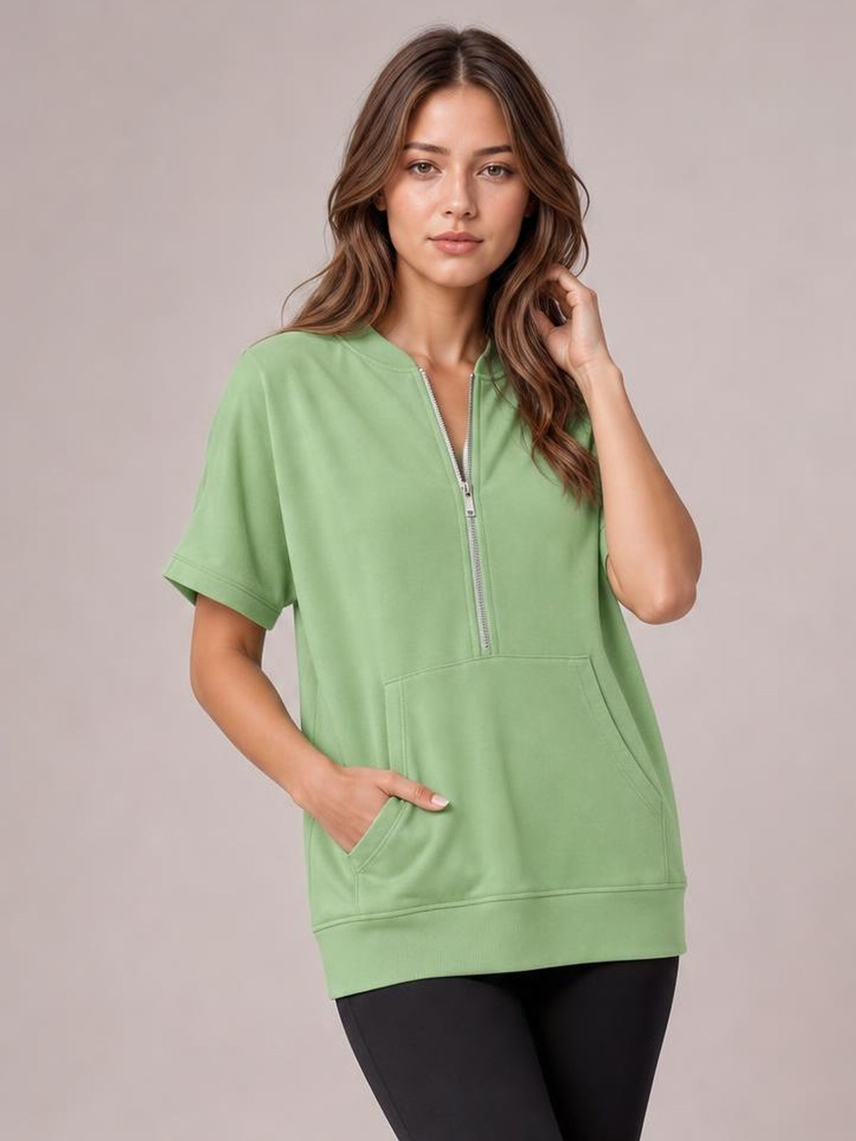 Model wearing a sage green half-zip short-sleeve hooded top with a relaxed long silhouette, visible seam details, and a large kangaroo pocket.
