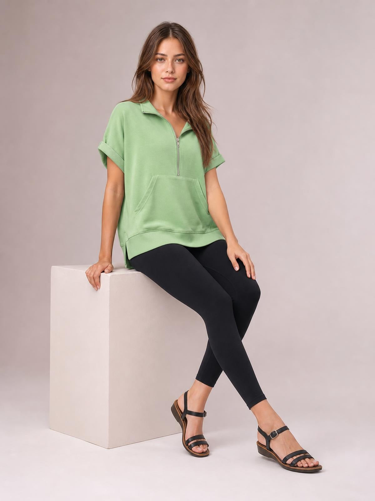 A seated model wears a sage green half-zip short-sleeve top with a relaxed silhouette, ribbed cuffs and hem, and a spacious front pocket.