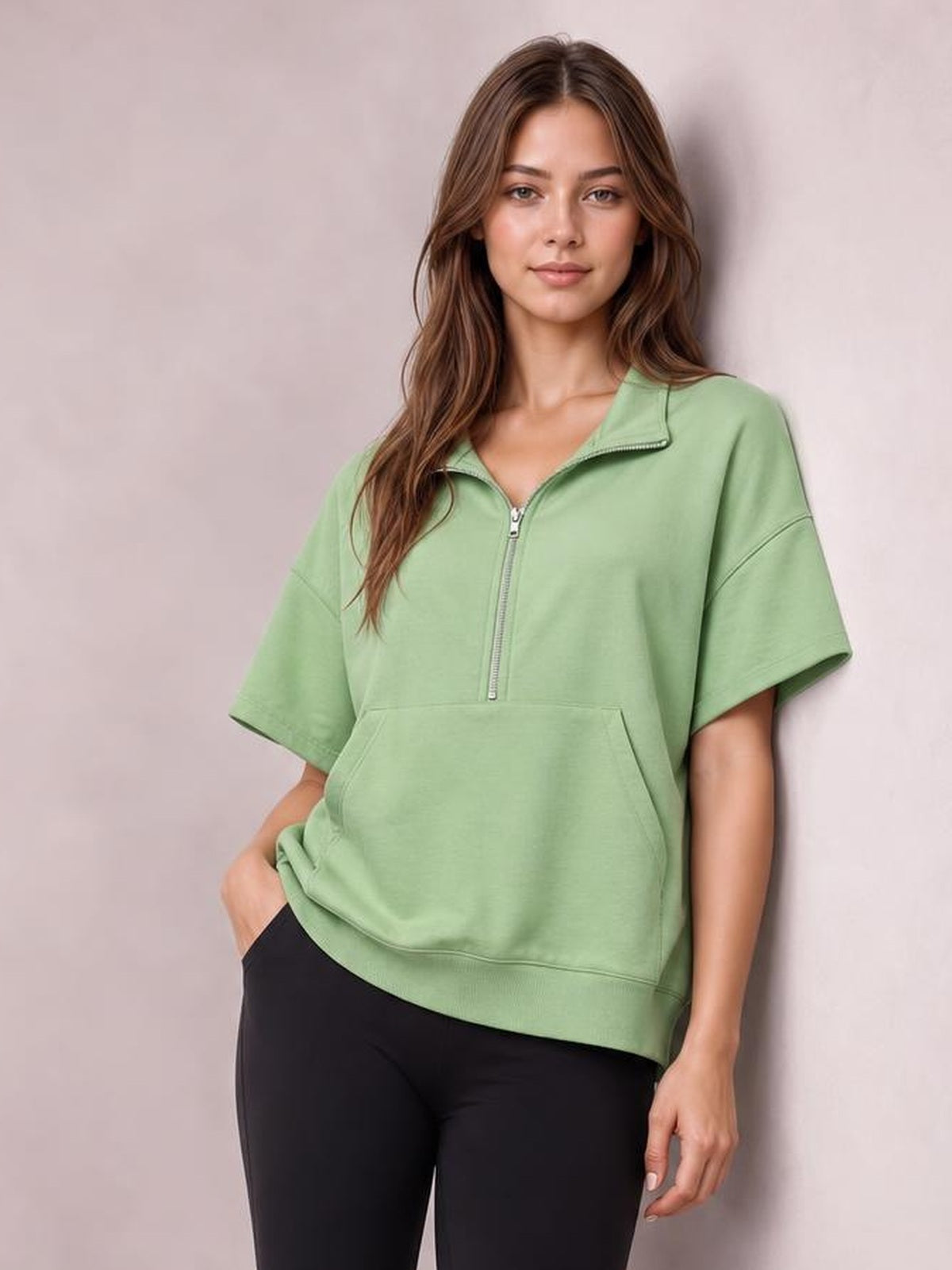 Model wearing a sage green half-zip short sleeve hooded top with a relaxed silhouette, stand collar, kangaroo pocket, and ribbed hem.