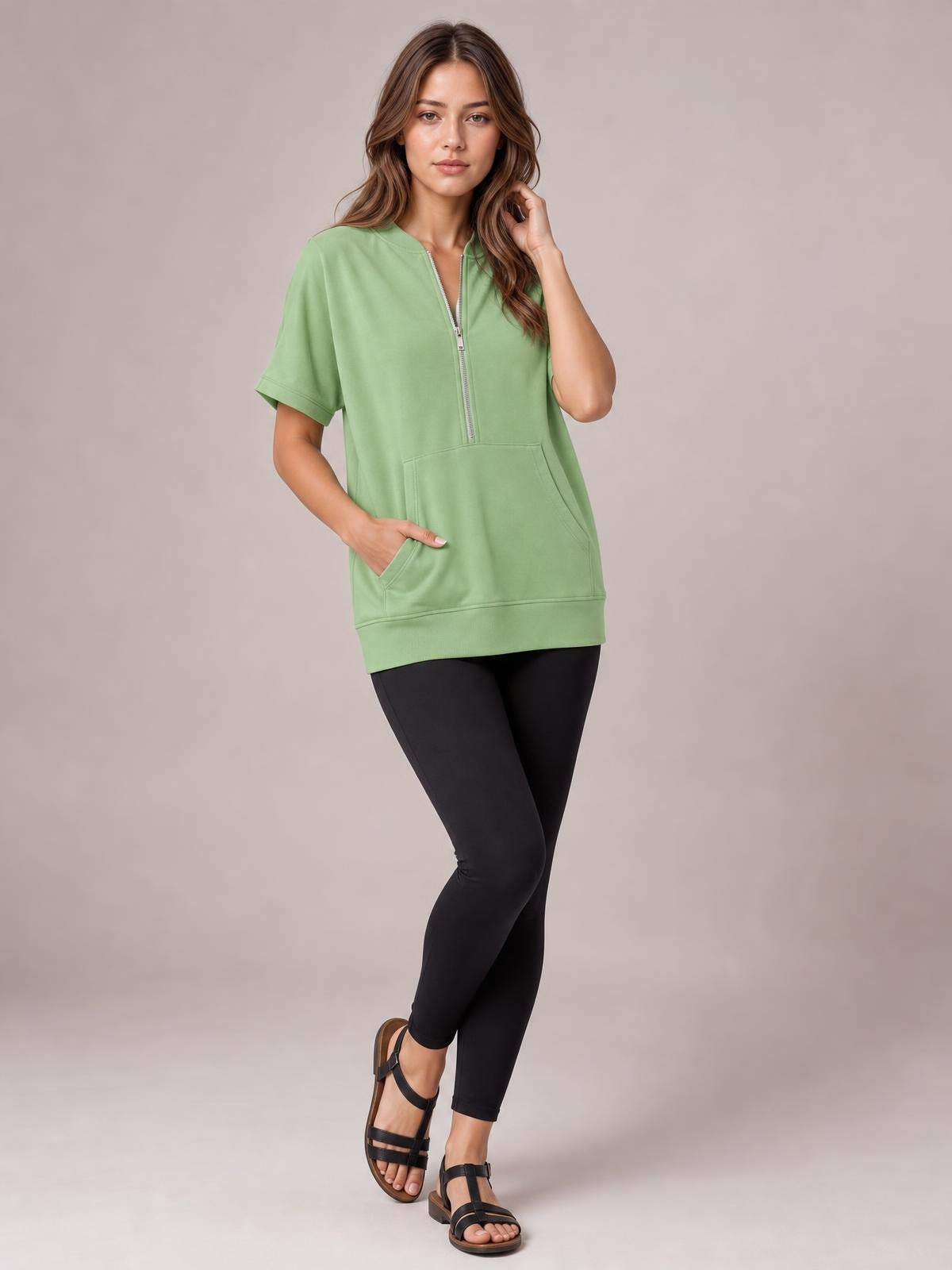 Model poses in a sage green half-zip short-sleeve hooded top with a relaxed tunic silhouette, ribbed hem, and front pouch pocket.