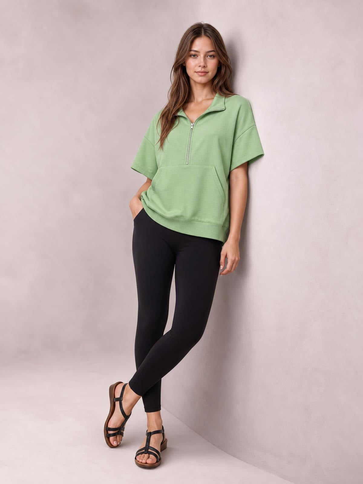 A model poses in a relaxed sage green half-zip short-sleeve hooded top with a roomy silhouette, front pocket, and ribbed hem.