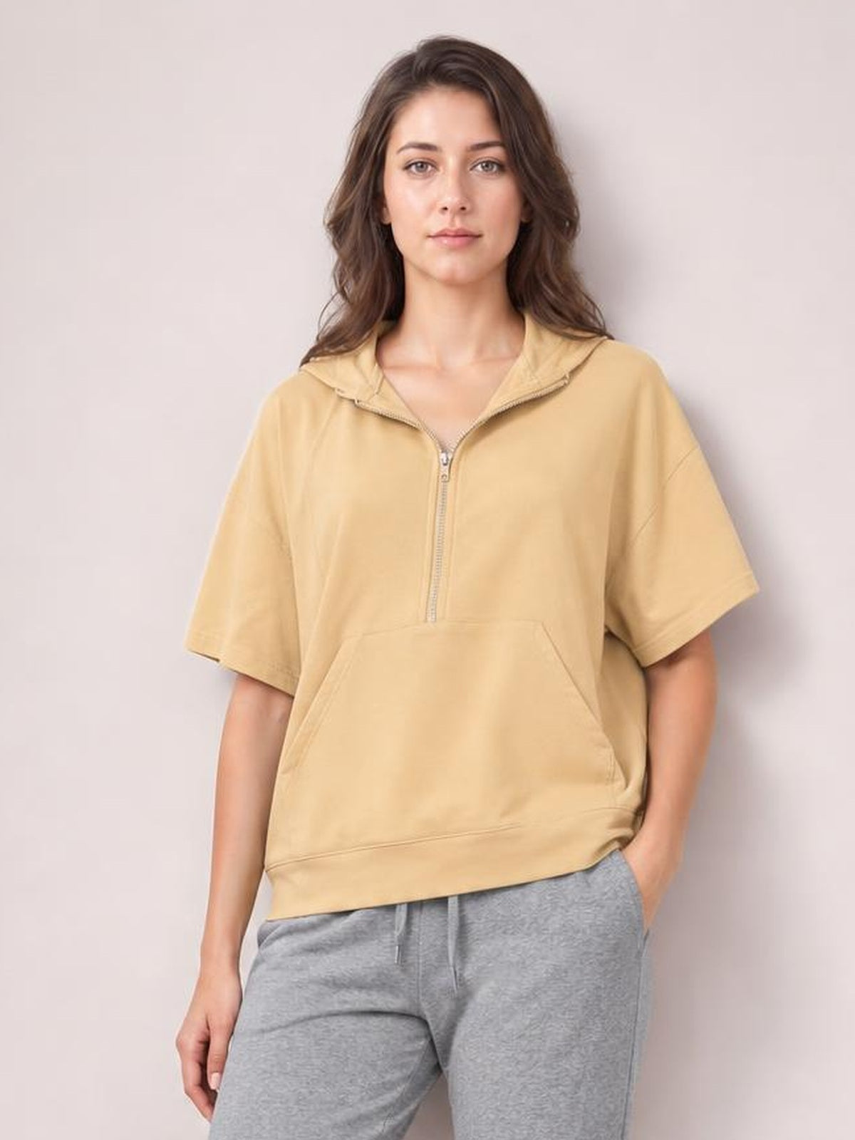 Model wears a relaxed tan half-zip short-sleeve hooded top with a front kangaroo pocket, visible zipper, and softly structured silhouette.