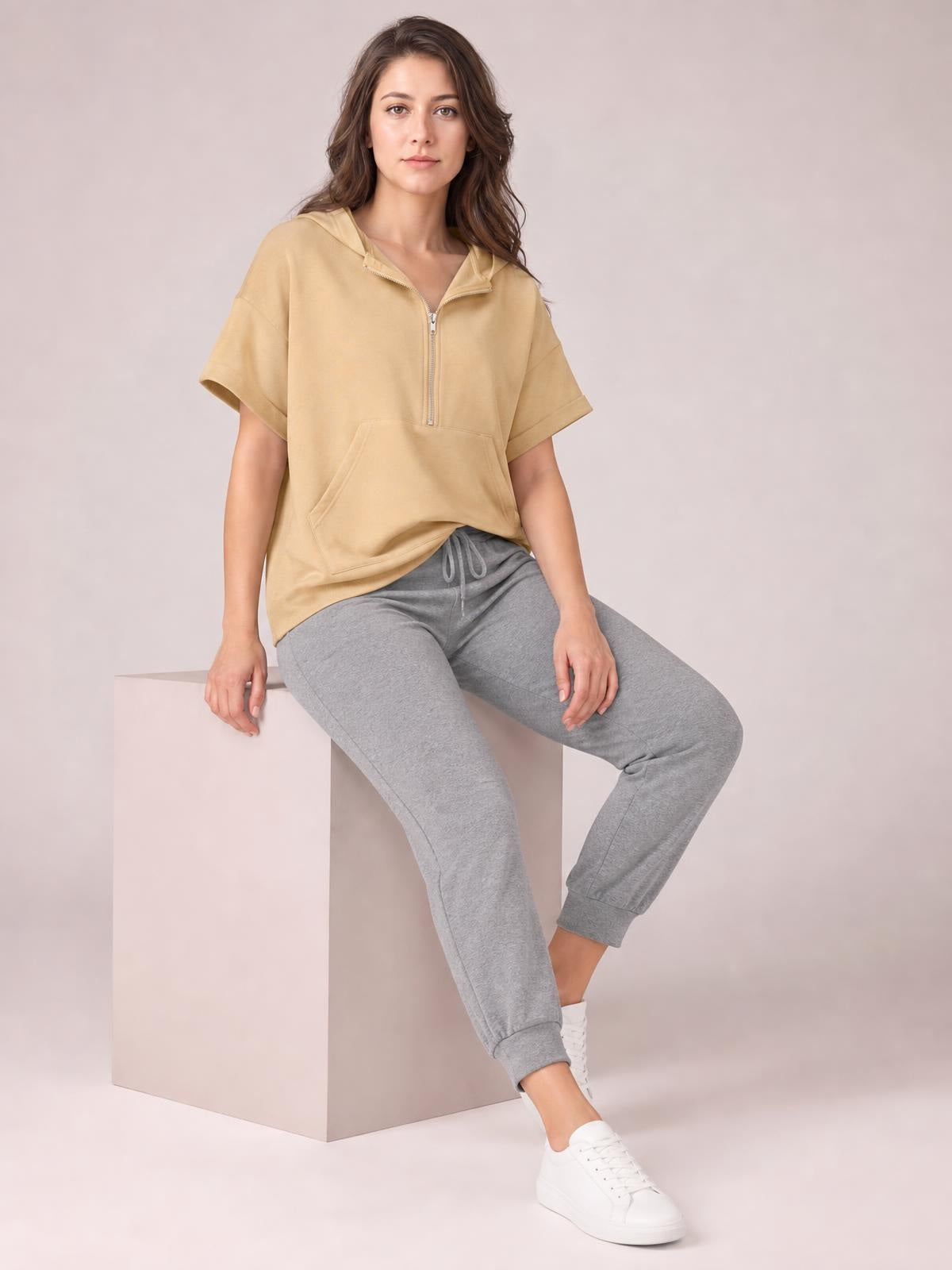 A woman sits wearing a tan short-sleeve hooded top with a half-zip front, relaxed silhouette, and spacious kangaroo pocket.