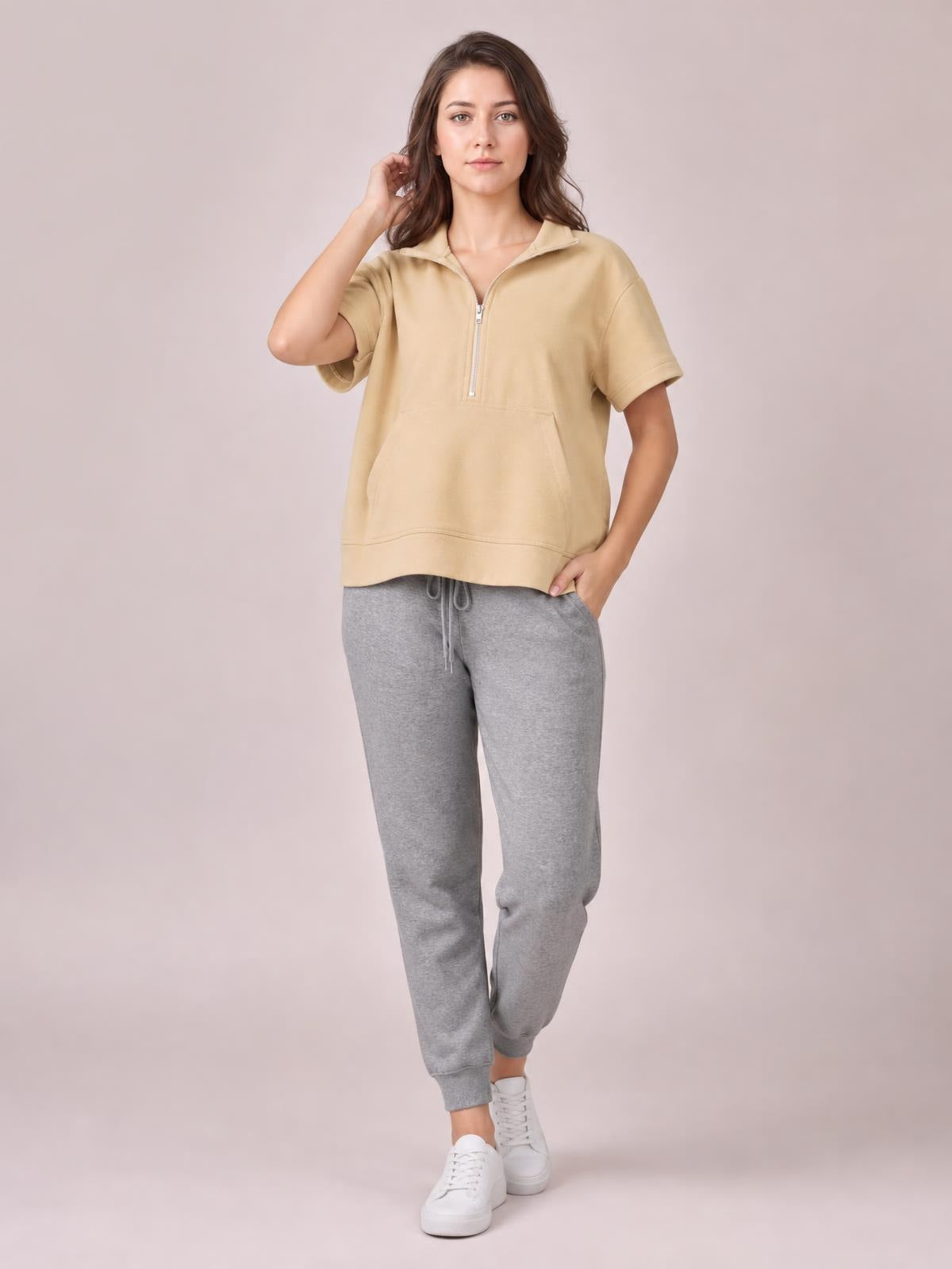 Model wears a tan half-zip short-sleeve top with a structured collar, relaxed silhouette, and large front pocket, posing with one hand in her pocket.