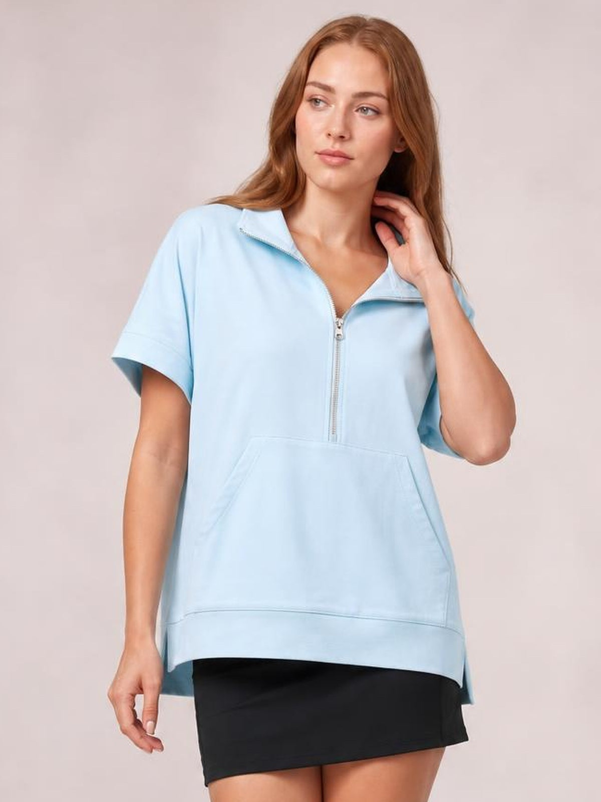 A model poses in a light blue short-sleeve hooded top with a half zip, relaxed long silhouette, and roomy kangaroo pocket, touching her hair.