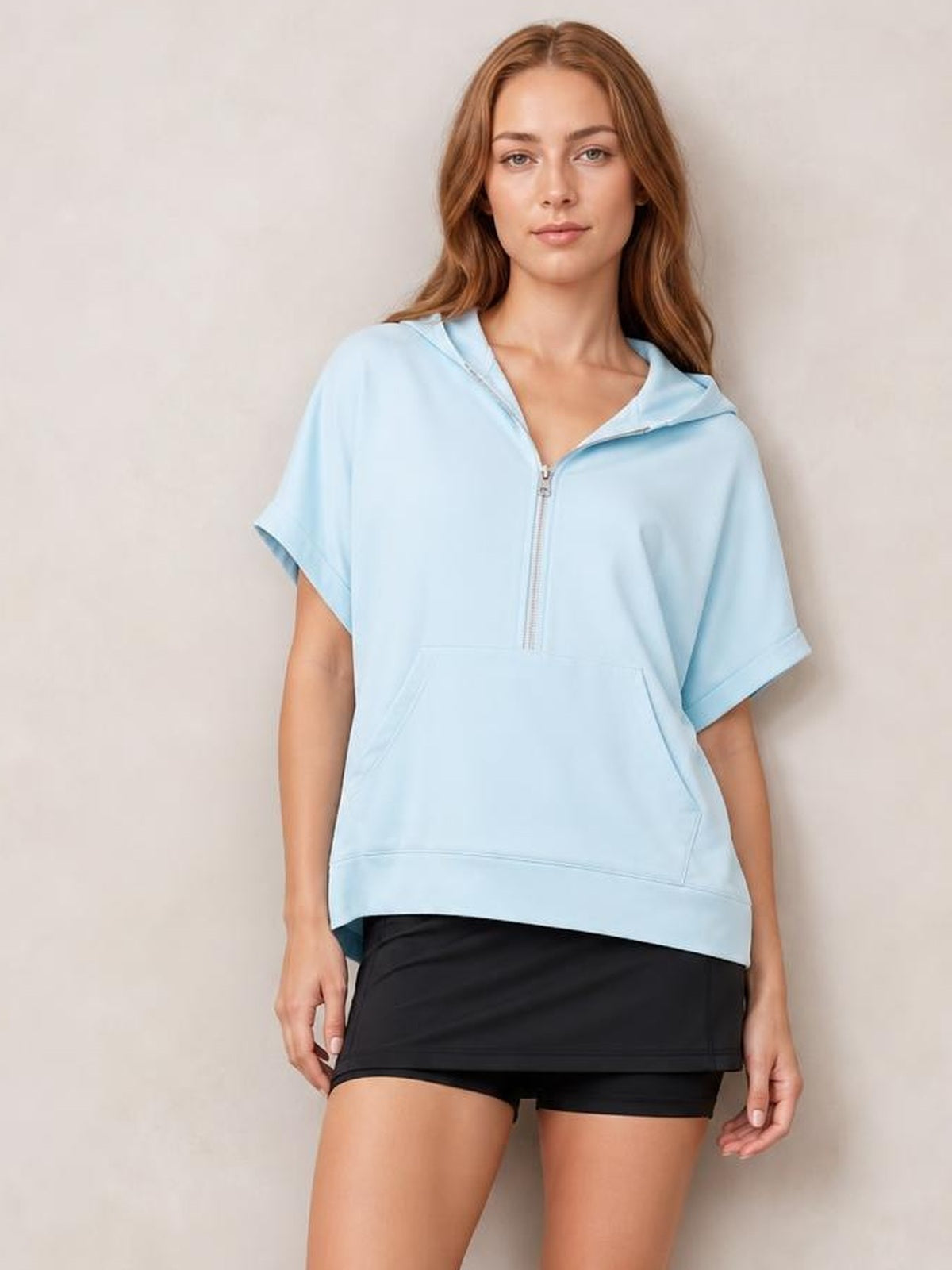 Model wears a light blue short-sleeve hooded top with a half-zip front, relaxed oversized silhouette, and large kangaroo pocket.