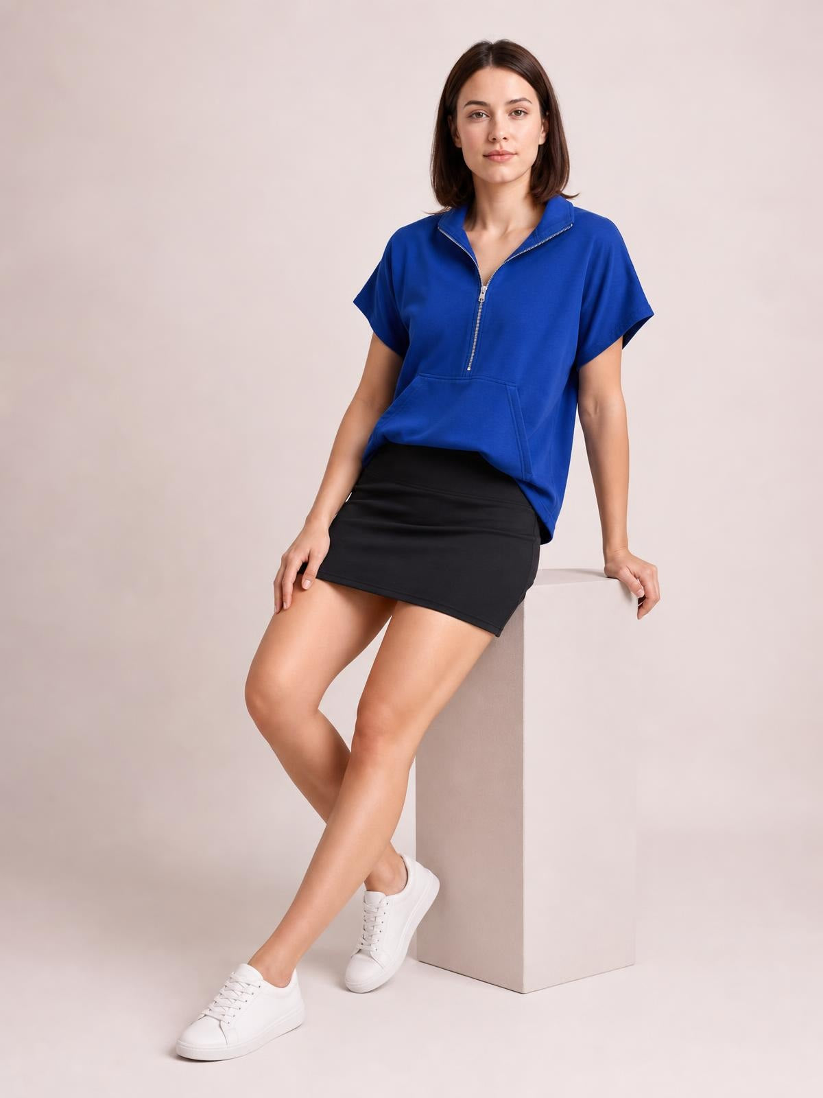 Model in a seated pose wears a royal blue short-sleeve hooded top with a half-zip front, relaxed silhouette, and large front pocket.
