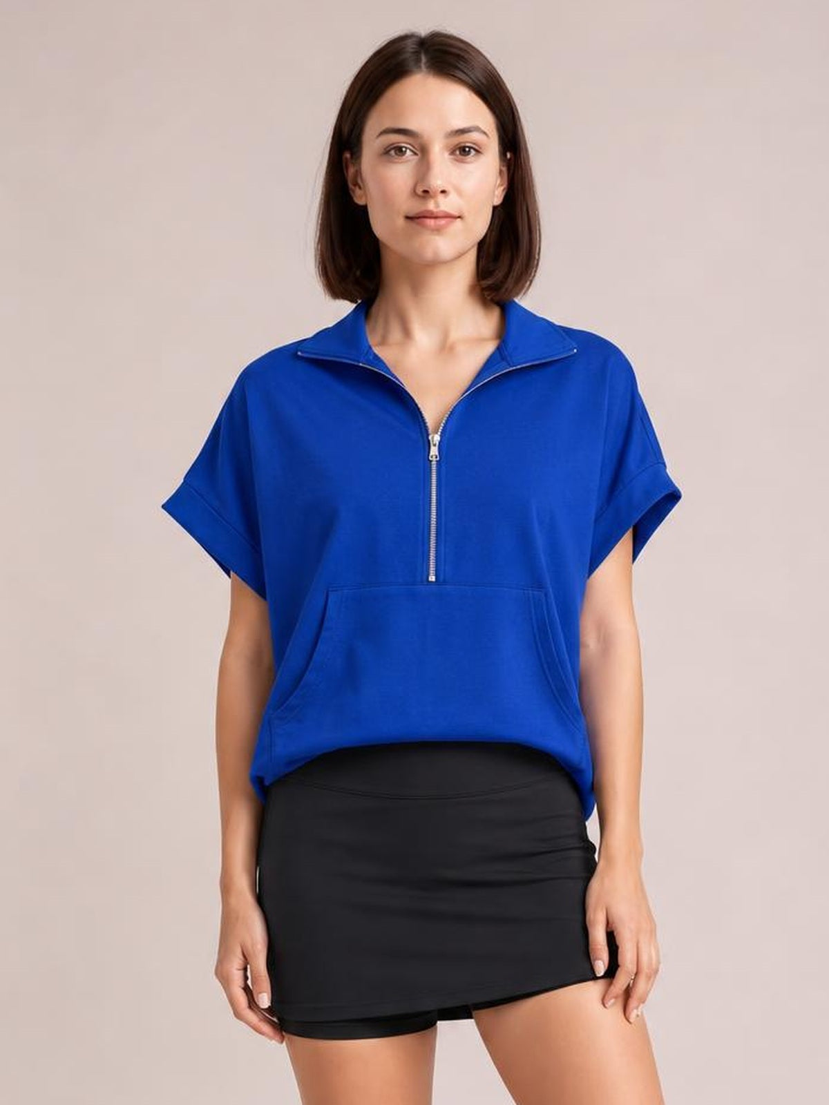 Model stands facing forward in a bright blue short-sleeve hooded top with a half-zip front, relaxed silhouette, and roomy kangaroo pocket.