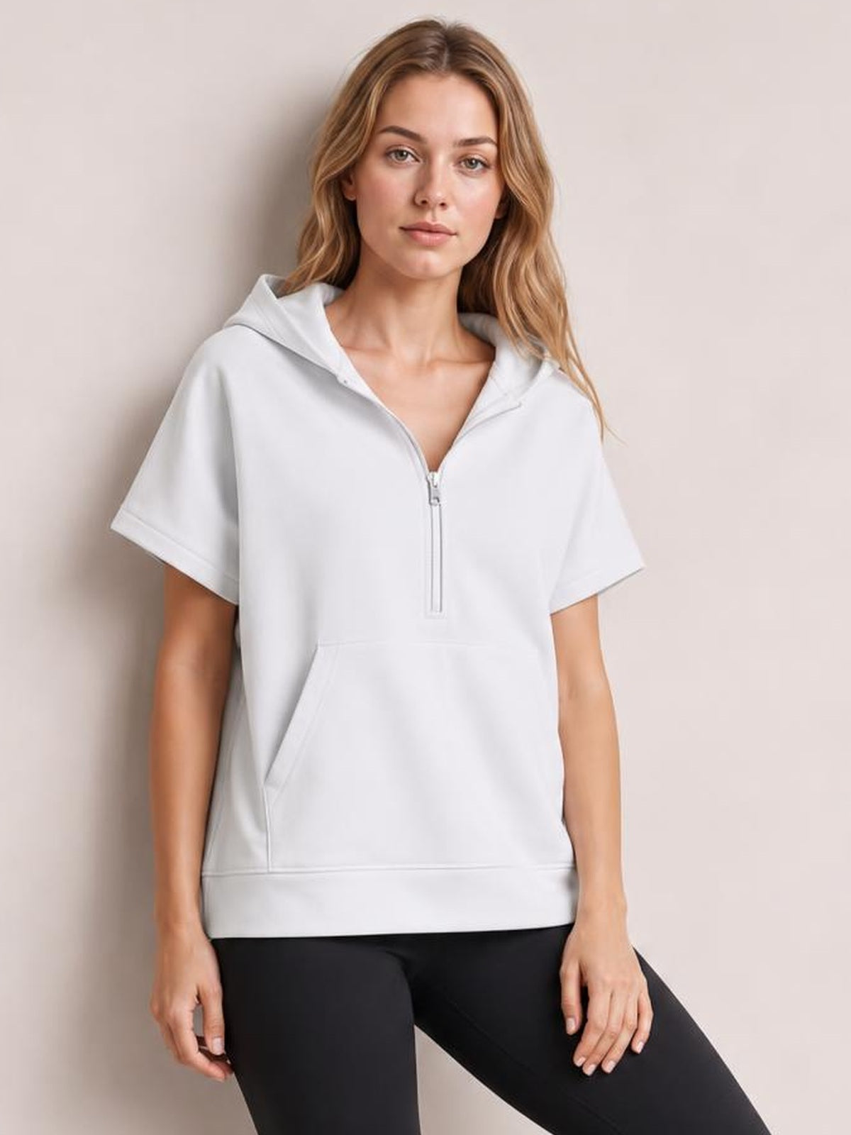Model stands wearing a white short-sleeve hooded top with a half-zip neckline, relaxed silhouette, and front kangaroo pocket.