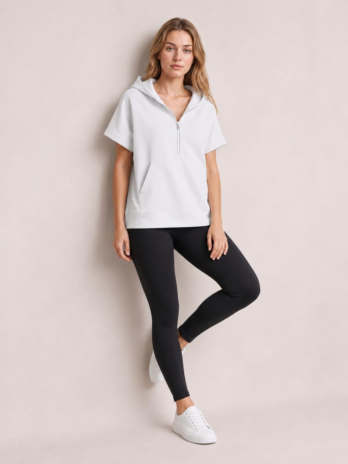 Model poses in a white short-sleeve hooded top with a half-zip front, kangaroo pocket, relaxed silhouette, and ribbed hem over black leggings.