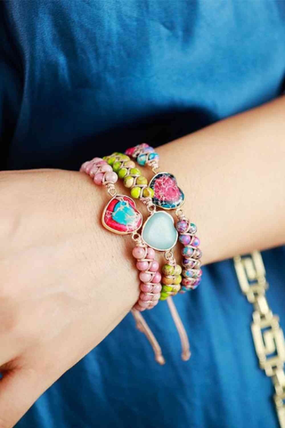 A model’s wrist displays a handmade heart shape natural stone bracelet with colorful beads, copper links, and a layered modern silhouette.
