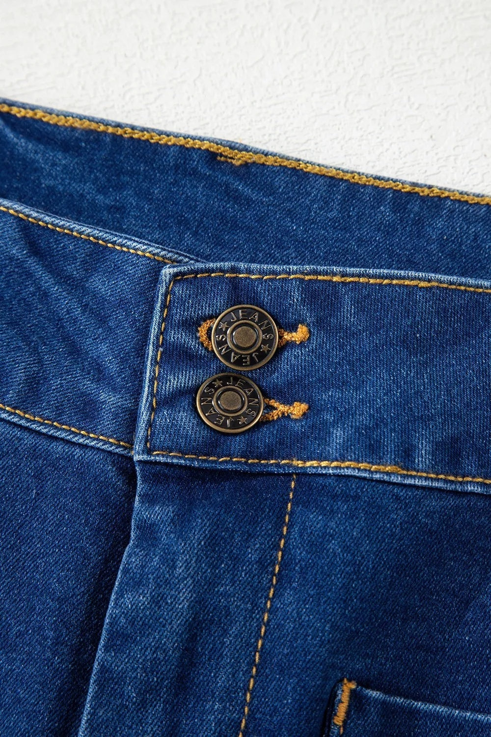 Close-up of dark blue wide-leg jeans showing a buttoned waistband, metal hardware buttons, patched pockets, and contrasting gold stitching.