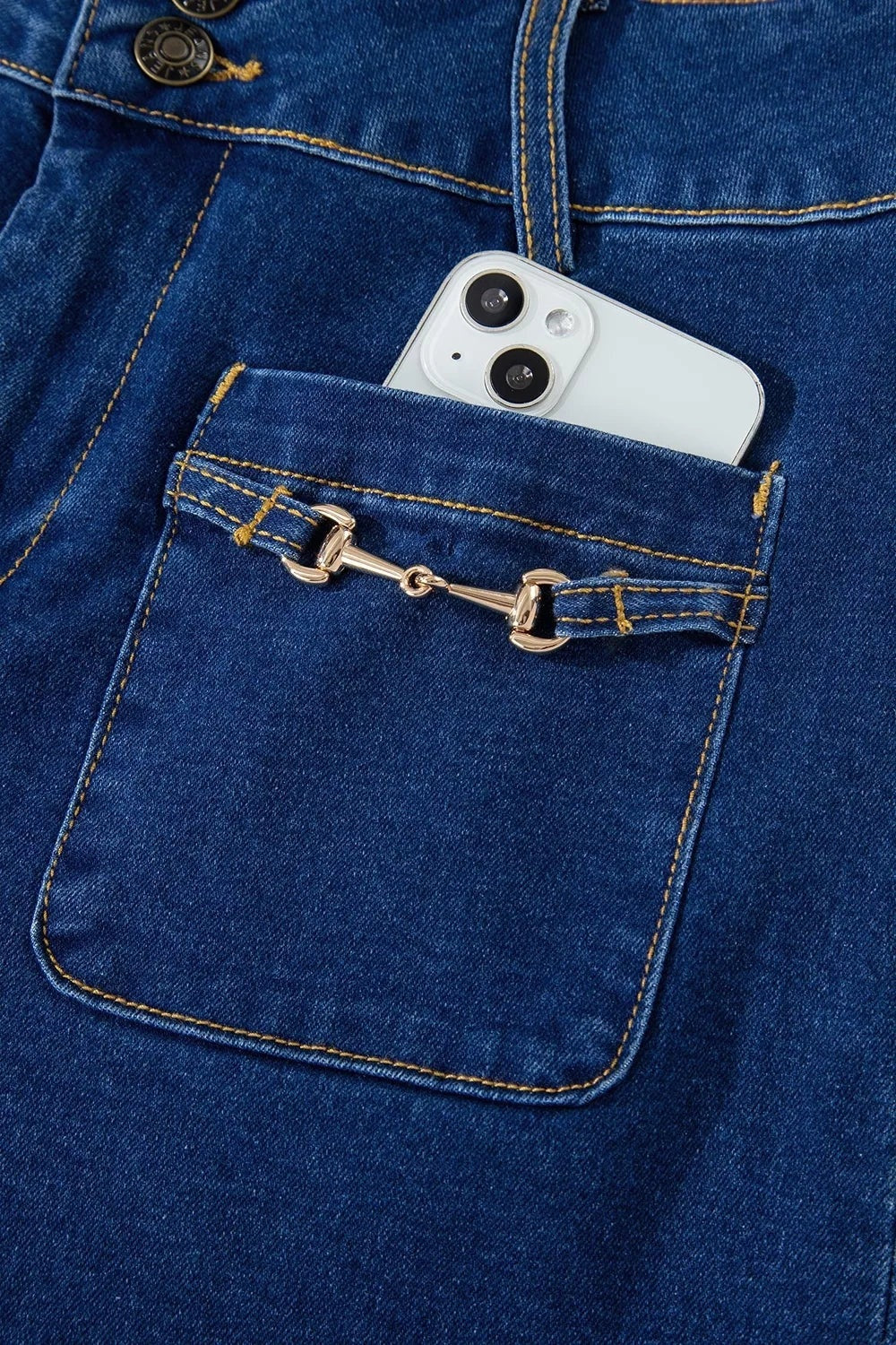 Dark blue jeans feature a large patched pocket with rounded edges, contrast stitching, button closure, and decorative gold-tone hardware.