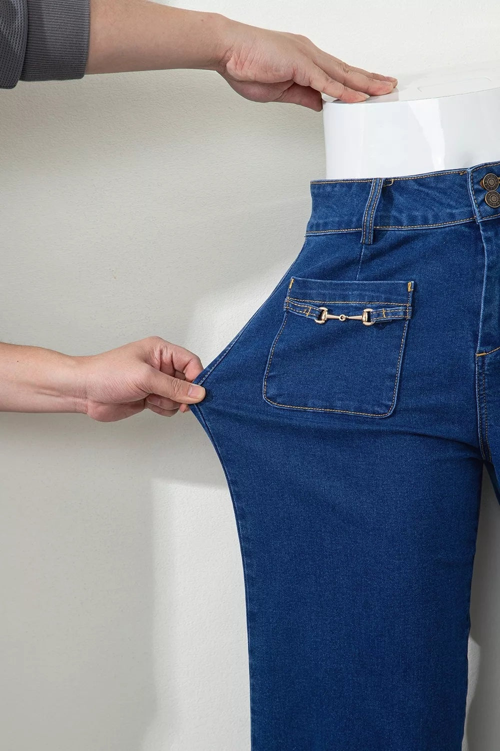 Close-up of dark blue wide-leg jeans showing a high-rise waistband, button closure, patched pocket, contrast stitching, and decorative hardware detail.