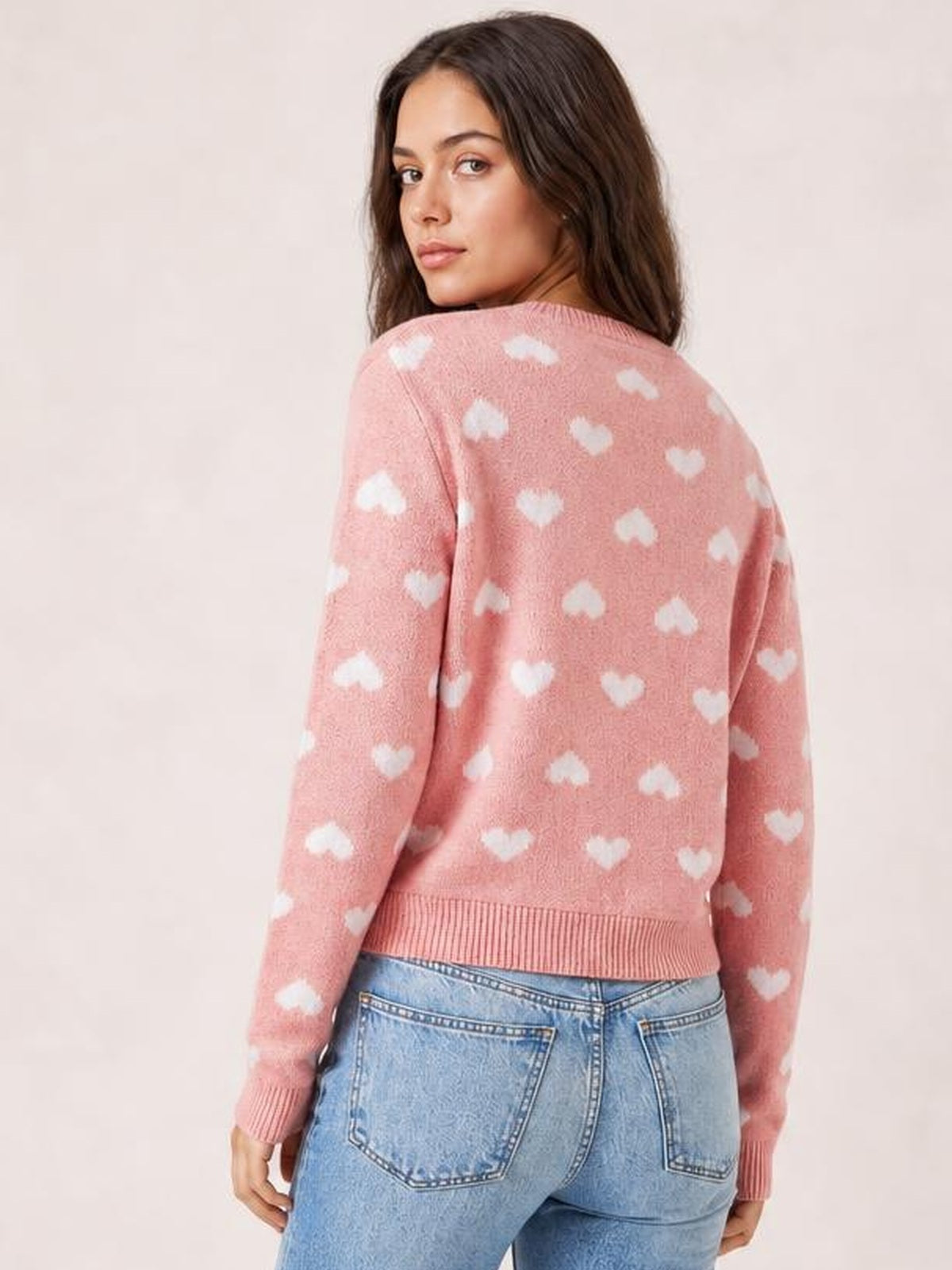 Model looks over her shoulder wearing a pink heart-pattern cardigan with long sleeves, a relaxed silhouette, and ribbed cuffs and hem.