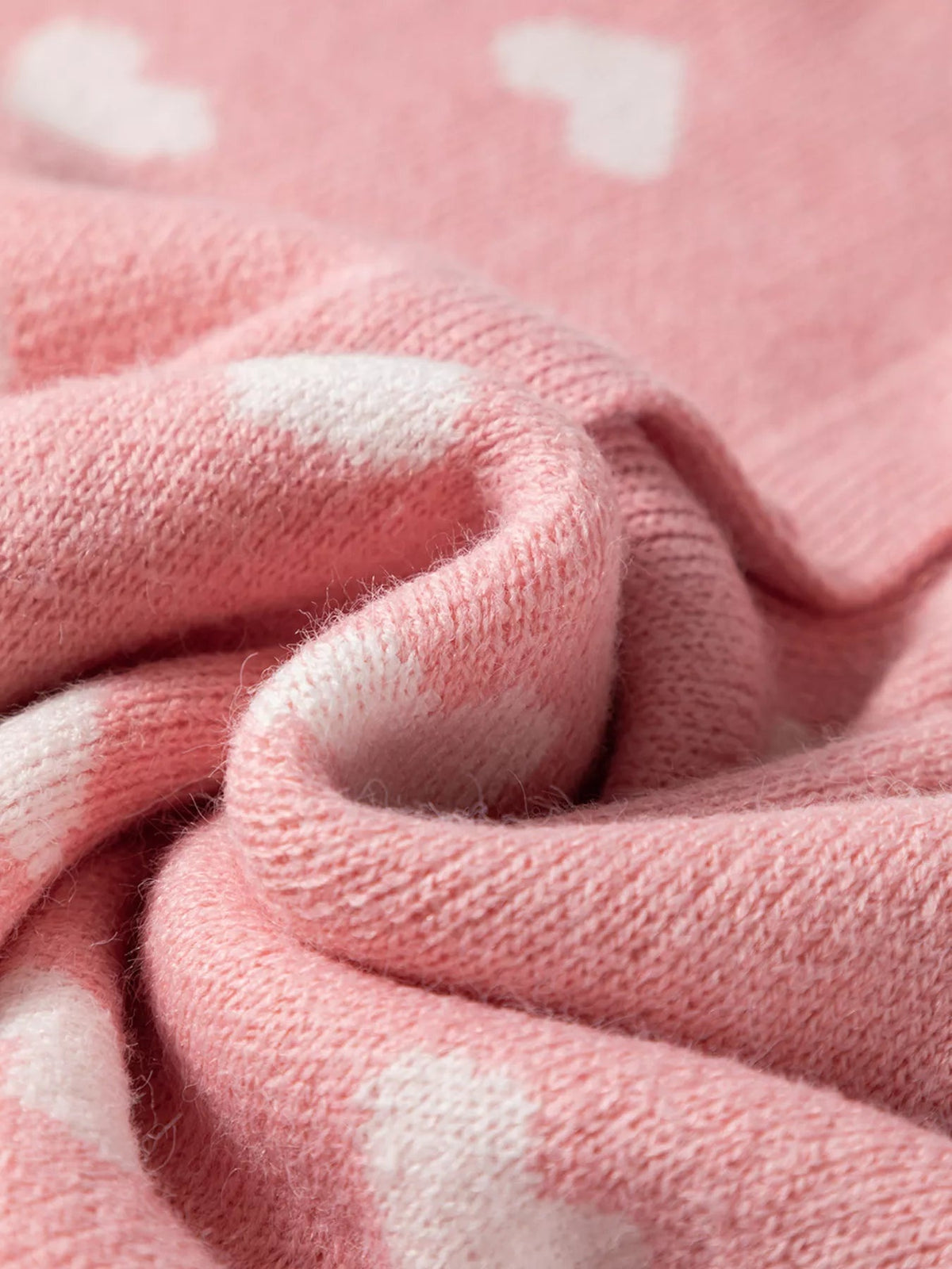 Close-up of a soft pink knit cardigan with a white heart pattern, showing its fuzzy texture and gently folded, slightly stretchy construction.