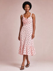 Model stands with crossed legs in a pink heart-print cowl neck midi dress featuring ruched draping, thin straps, a fitted waist, and a ruffled hem.