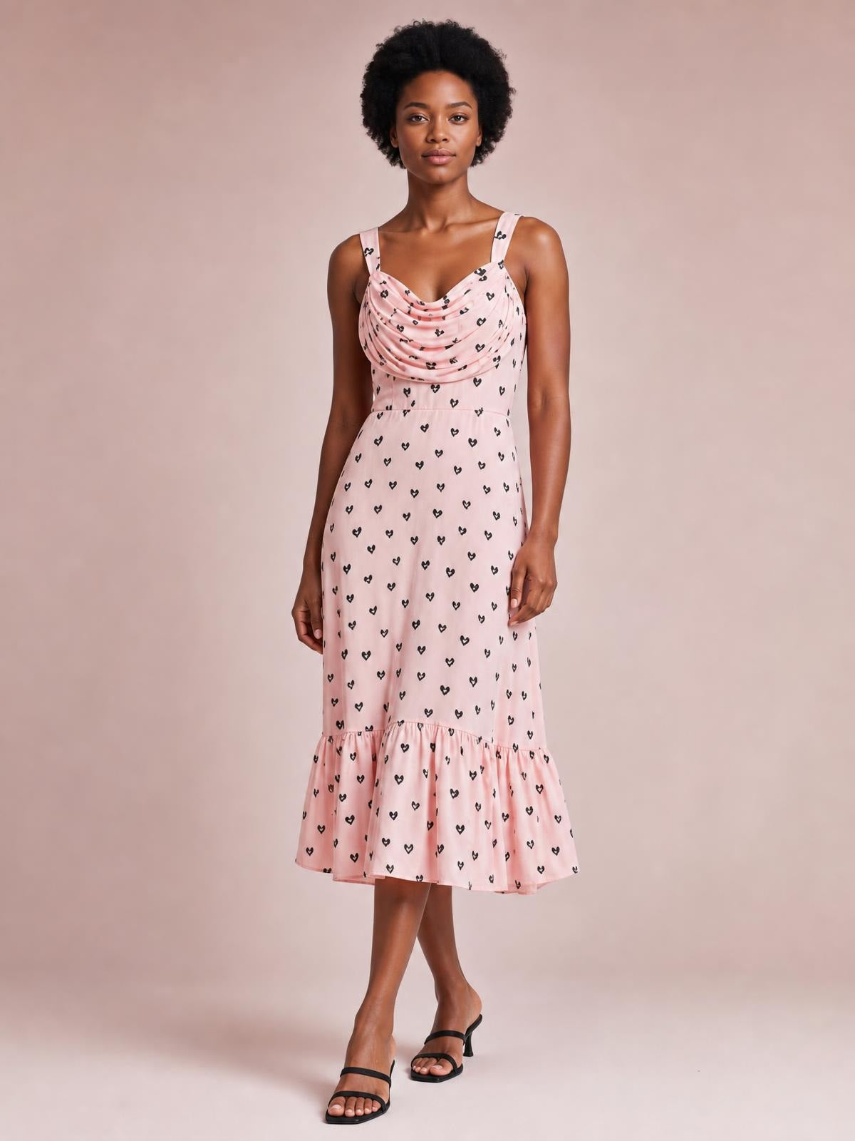 Model stands with crossed legs in a pink heart-print cowl neck midi dress featuring ruched draping, thin straps, a fitted waist, and a ruffled hem.
