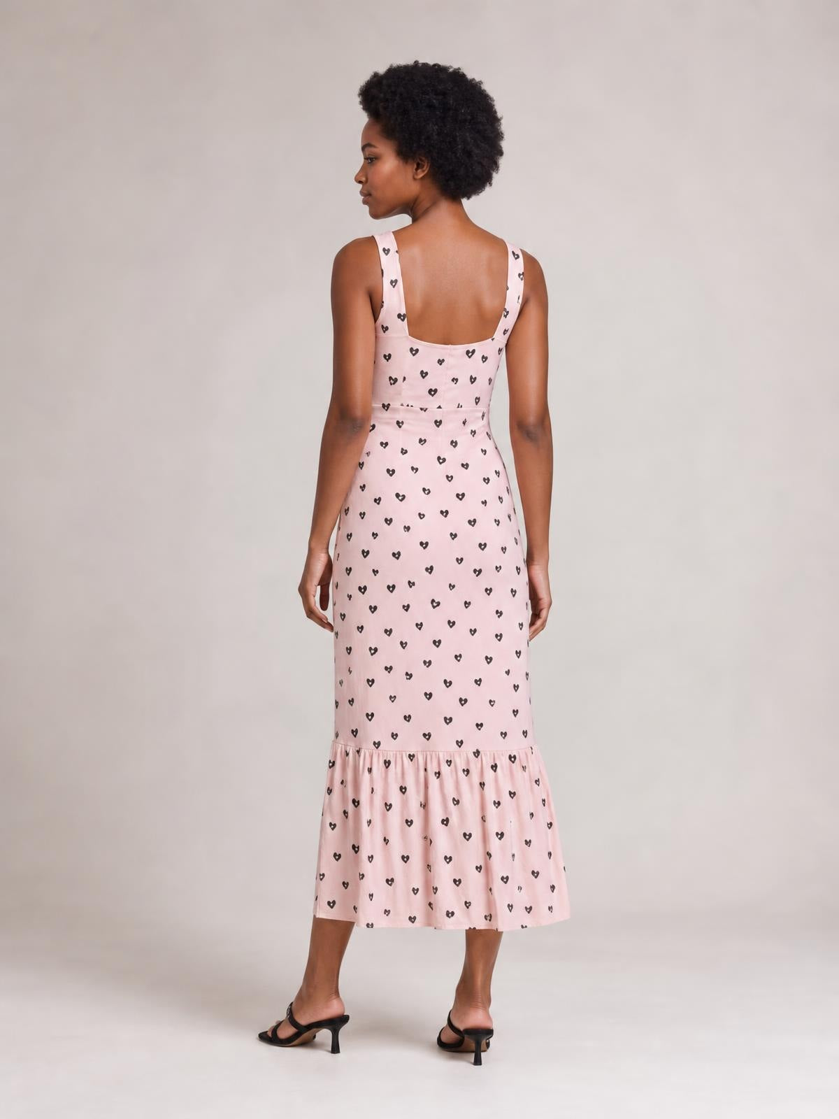 A model shows the back of a pink heart-print midi dress with wide straps, a fitted ruched waist, straight silhouette, and ruffled hem.