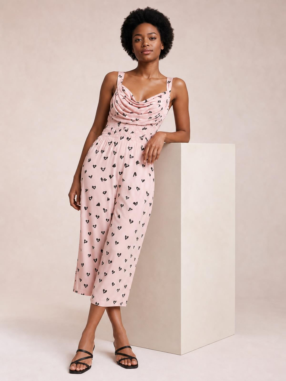 Model wears a pink heart-print cowl-neck jumpsuit with shoulder straps, a ruched waist, and a cropped wide-leg silhouette.