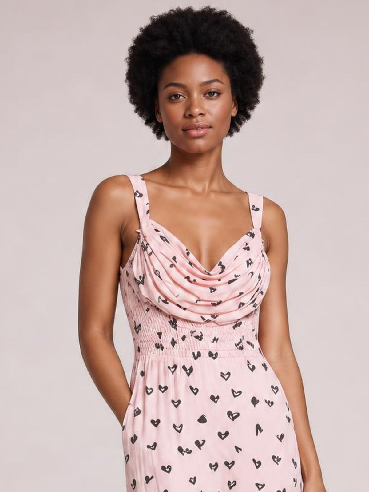 Model wearing a pink heart-print midi dress with a draped cowl neck, wide straps, ruched waist, and softly flowing A-line silhouette.
