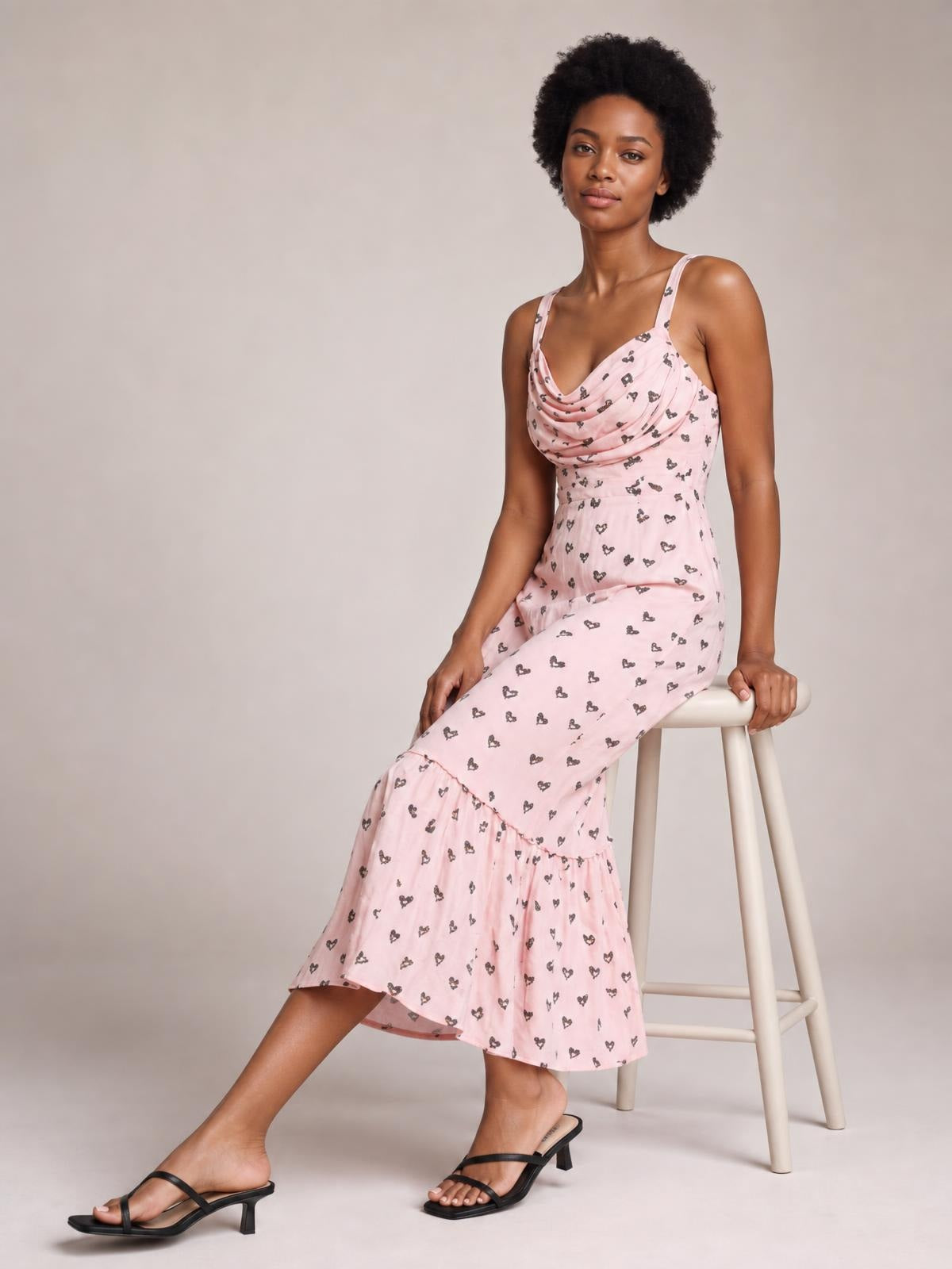 A seated model wears a pink heart-print midi dress with a draped cowl neck, thin straps, fitted ruched bodice, and ruffled flared hem.
